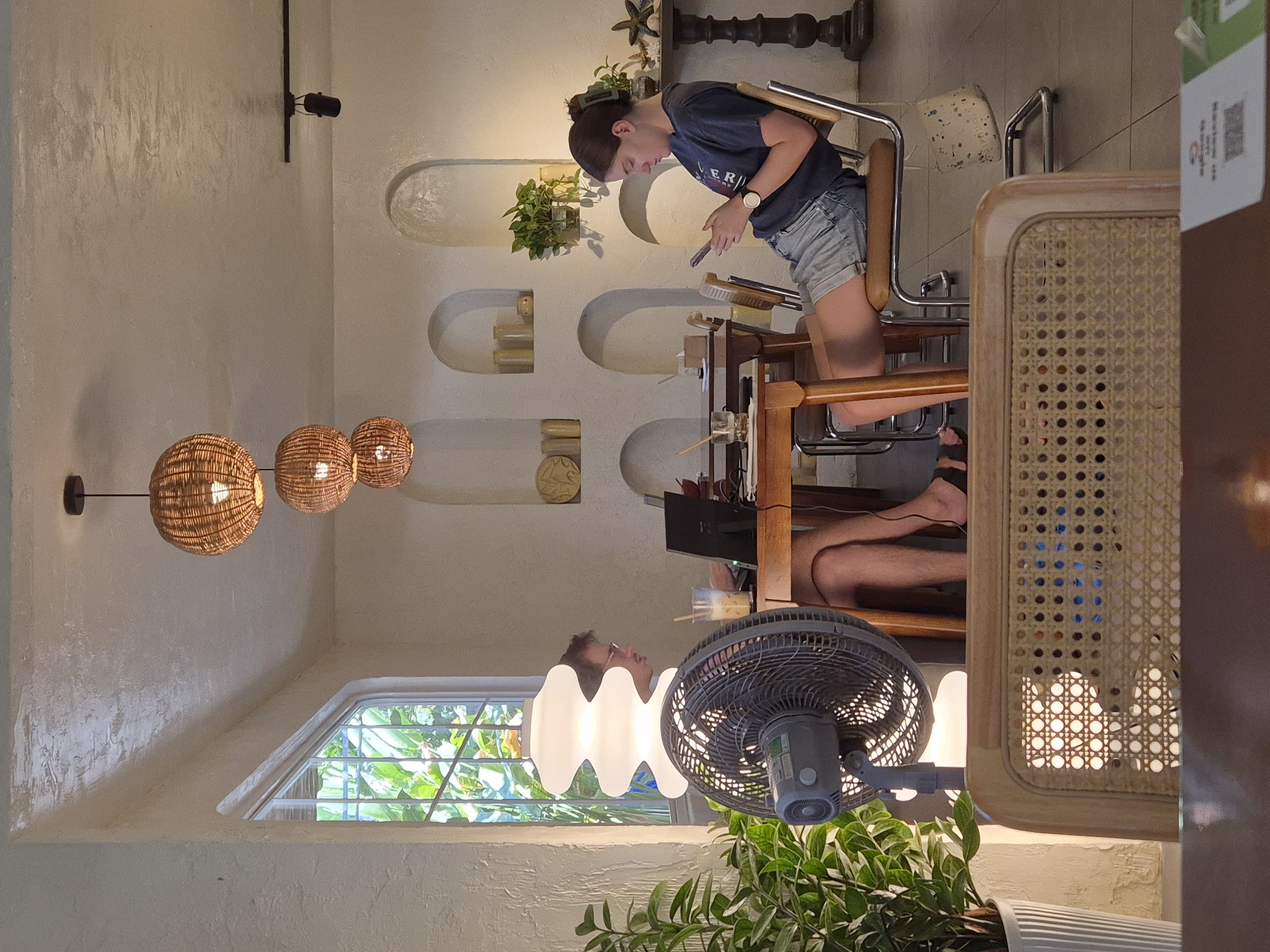 In a brightly lit interior space, possibly a cafe in Đà Nẵng, Vietnam, two individuals are seated at a wooden table. On the left, a man with glasses is engrossed in his laptop, with a tall, layered white lamp and a large fan positioned nearby. In front of him, a glass with a straw and ice sits on the table. To his right, a woman is looking down at her mobile phone, wearing a dark t-shirt and denim shorts. Her chair is made of metal and has a leather seat, and a small patterned bag hangs from it. The room features a textured white wall with arched niches, some containing decorative items and plants. Natural light streams in from a window on the left, revealing lush greenery outside. Three woven pendant lamps hang from the ceiling, casting a warm glow. The flooring is tiled.