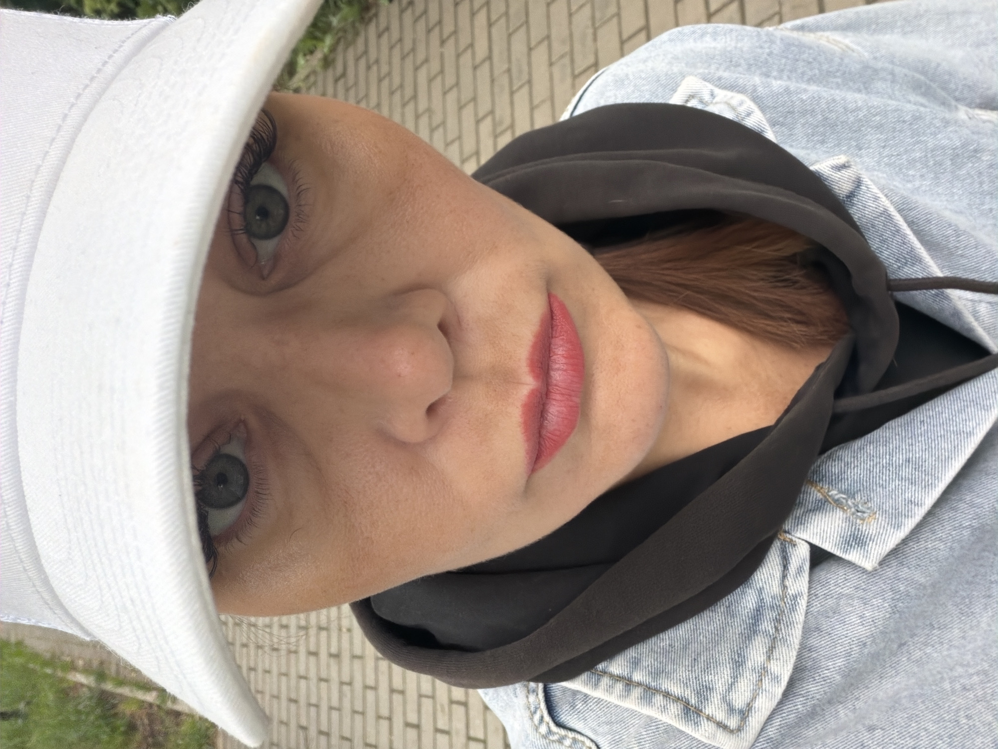 A close-up, low-angle selfie shows a woman from the chest up. She is wearing a white baseball cap, a black hoodie, and a light-wash denim jacket. Her face is tilted downwards, and her eyes are looking directly at the camera. The background consists of a paved walkway with brick-like pavers and some greenery on the left. The image appears to have been taken outdoors. The exact location in the city of Chaikovsky, Russia, cannot be determined from the visual information provided.