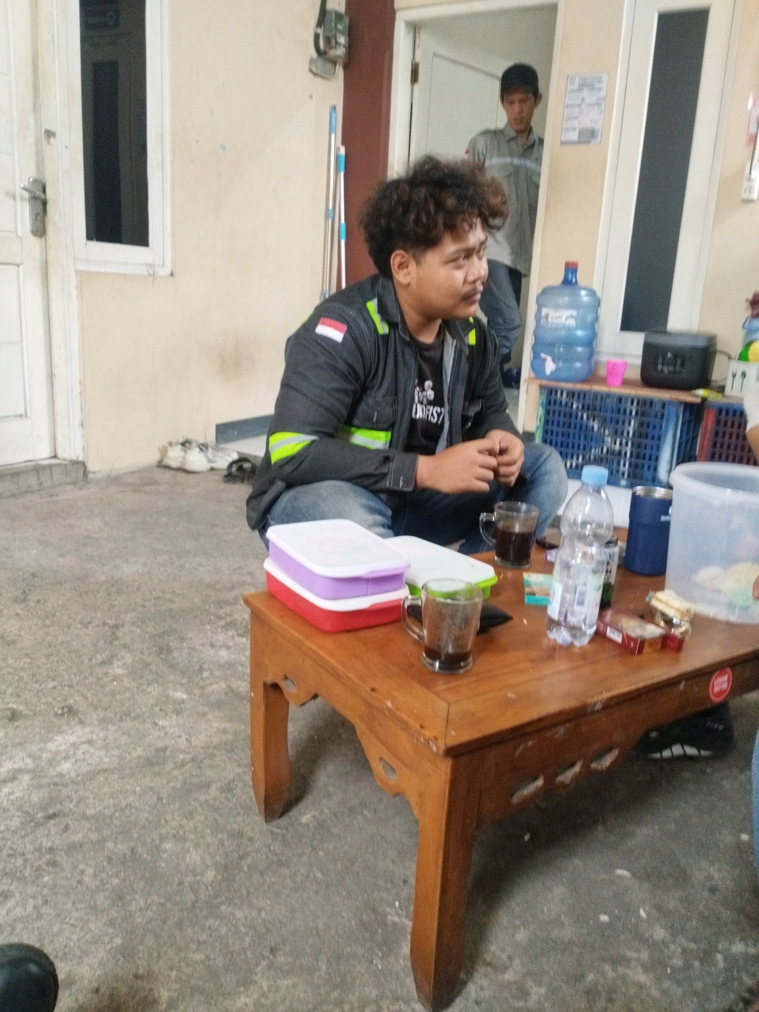 The image depicts an outdoor scene in Selomerto, Indonesia, likely in the vicinity of a residential or communal area. In the foreground, a wooden coffee table is laden with various items. Two glasses of dark liquid, likely coffee or tea, are present. A bottle of water stands next to them, along with a pack of cigarettes and some unwrapped candy. Two rectangular food containers, one red and one purple with a white lid, are stacked on the table. A person with curly dark hair, wearing a dark jacket with reflective green and yellow stripes and blue jeans, is seated on the ground, facing the table. Their posture suggests they are engaged in conversation or enjoying a break.
In the background, a large blue water cooler jug is visible, positioned on a blue slatted shelf unit. Adjacent to it is a small black electronic device. A second individual, a man wearing a grey shirt and a dark baseball cap, stands in a doorway behind the first person. The environment appears to be an open, paved area, possibly a patio or walkway, with a building wall and a door visible. A white window is set into the wall to the left. Scattered on the ground near the wall are a pair of shoes. The overall impression is of a casual gathering or a pause in activity.