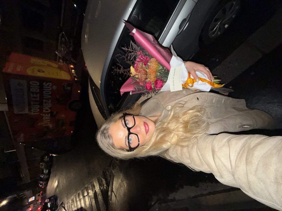A woman with blonde hair and glasses poses for a selfie at night. She is holding a bouquet of flowers wrapped in pink paper and tied with a yellow ribbon. A white car is visible behind the flowers. A delivery truck with "TOUT LE BIO LIVRÉ CHEZ VOUS" written on its side is parked on the street in the background, amidst other parked cars. The street appears to be wet, reflecting the lights of the city.