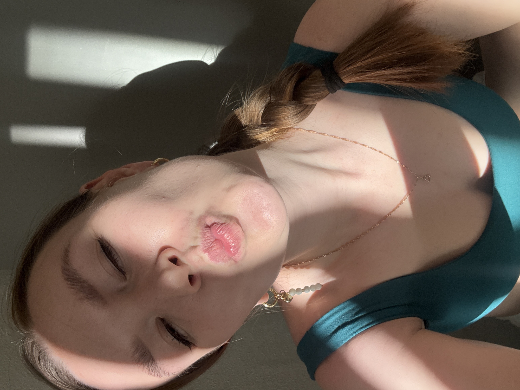 An angled, close-up photograph captures a person lying down indoors, illuminated by strong, direct sunlight. The image is rotated counter-clockwise, showing the person's head at the bottom left and their torso extending towards the top right. The subject is a young individual with light skin and light brown hair styled in a braid, secured by a black hair tie. Their eyes are closed, and their lips are puckered in a playful or affectionate pose.
They are wearing a dark teal or green top, possibly a swimsuit. Accessories include a delicate gold-colored chain necklace with a small, indistinct pendant, and another beaded necklace featuring light green or white beads and a circular charm with a small lock. A gold-toned earring is visible in the left ear.
Harsh shadows are cast across the plain, light-colored background and the person's body, indicating direct sunlight, likely from a window, suggesting either morning or late afternoon. The strong contrast creates distinct patterns of light and shade. The overall mood conveyed is casual and relaxed. There is no visible text. This scene is set in Izhevsk, Russia.