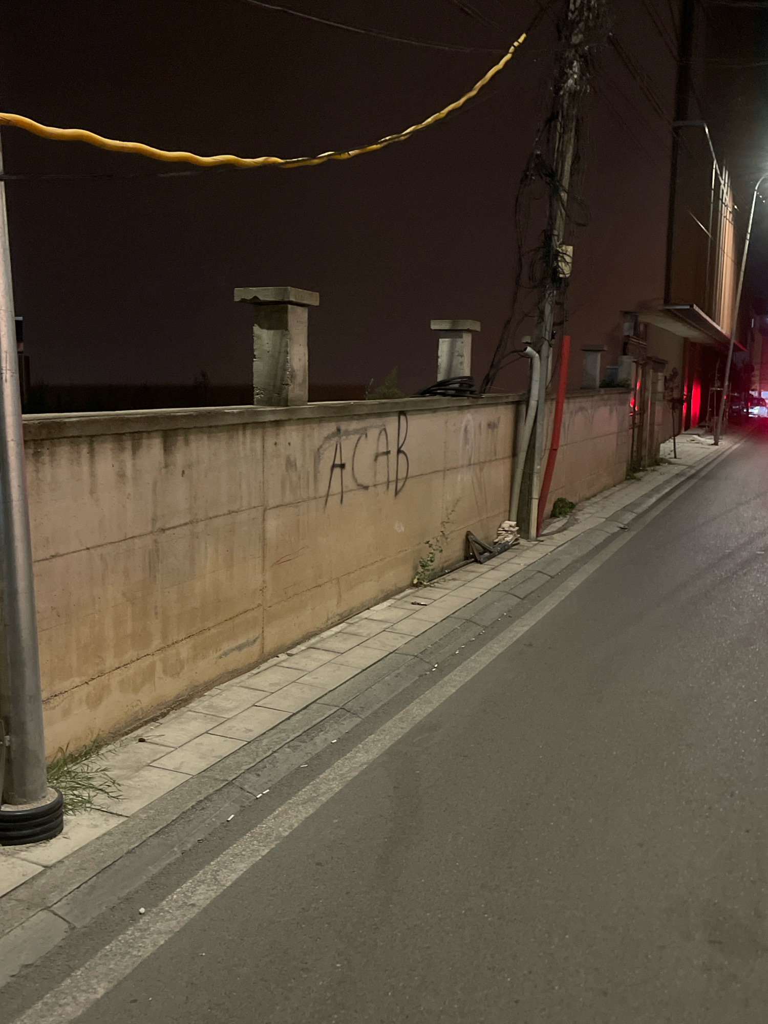 An outdoor, eye-level, medium shot captures a nighttime street scene in Prizren, Kosovo. The foreground is dominated by a grey asphalt road with a white stripe marking its edge. To the left of the road is a sidewalk paved with grey rectangular tiles, bordered by a curb. A grey metal pole with a black ribbed base stands on the left side of the sidewalk, partially obscuring the view. Beyond the sidewalk, a concrete wall stretches horizontally across the frame, its surface a pale beige with some staining. Graffiti in black, block lettering that reads "ACAB" is visible on the wall. Atop the wall, two square concrete structures are spaced apart, with another structure further in the background. Various pipes, including a red one, are attached to the wall and lean against it. In the background, a streetlamp illuminates a portion of the street to the right, and indistinct buildings and a dark sky are visible. The overall impression is one of an urban street at night, with evidence of graffiti and utilitarian infrastructure.