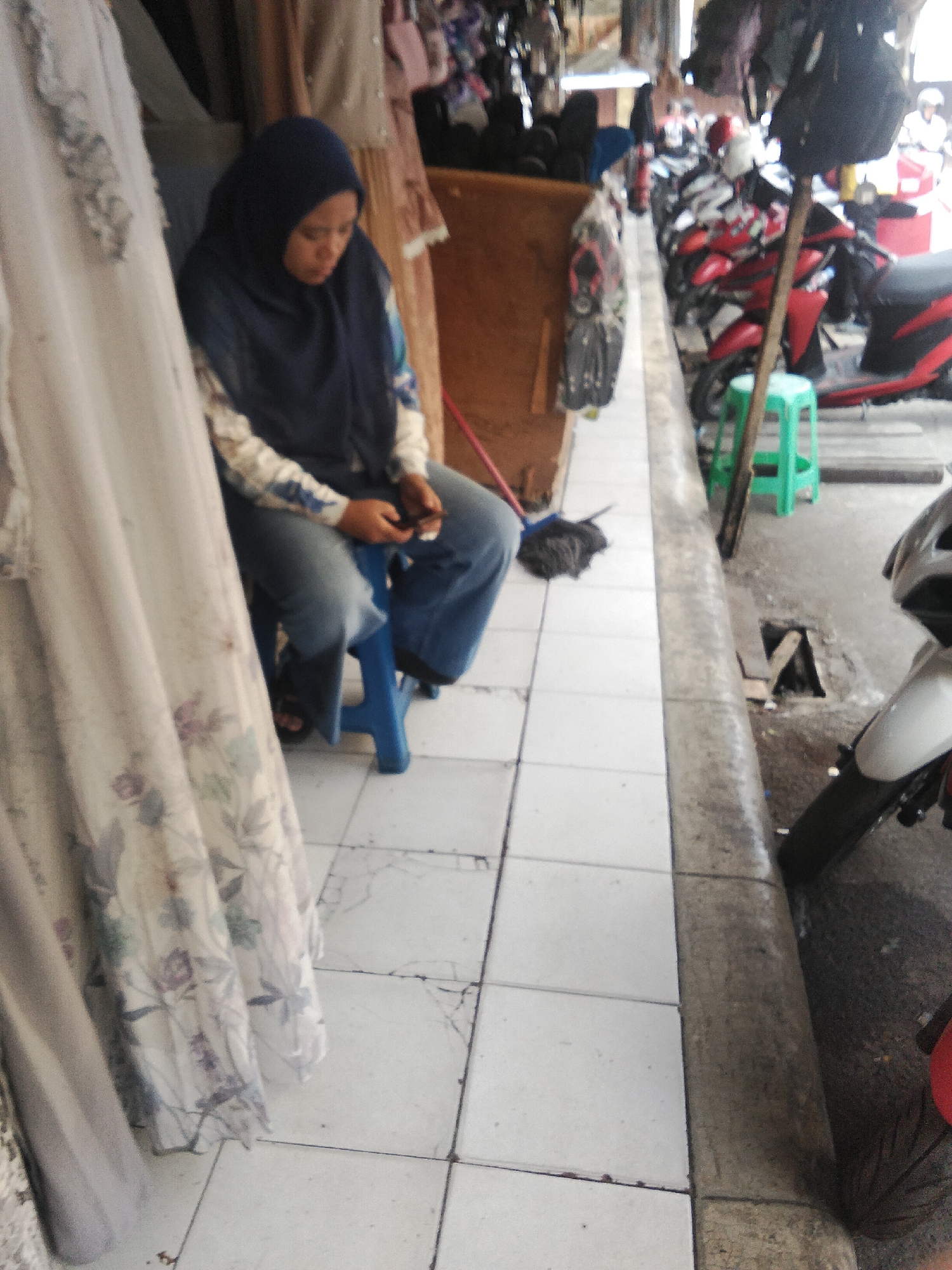 A woman wearing a dark blue hijab and a patterned long-sleeved shirt sits on a blue plastic stool in front of a stall selling clothing. She is looking down at a black mobile phone in her hands. She is wearing blue jeans and sandals. To her left, a floor-length garment with a floral pattern hangs on a display. In the background, several motorcycles are parked in a row along the street, and a green plastic stool is visible. The ground is tiled with white square tiles. The scene appears to be a street market in Cisaat, Indonesia.