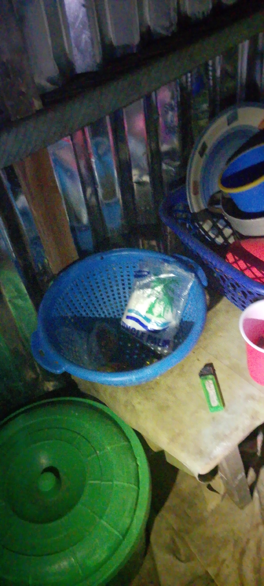 The image shows an indoor scene with various objects scattered on a wooden surface and the floor. In the foreground, a large green circular lid, possibly from a container, is visible on the left. To its right, a blue colander is placed on the wooden surface. Inside the colander, there appears to be a plastic bag containing white and green items, with text that includes "NICLE PALM." To the right of the colander, a green lighter lies on the wooden surface. Further to the right, a pink cup is partially visible, containing some dark liquid. In the background, stacked containers or bowls are visible, one with blue and yellow sections. The environment appears to be cluttered and somewhat disorganized. The lighting is dim, suggesting it might be nighttime or an indoor setting with poor illumination. There is no indication of the location in Jalingo, Nigeria.