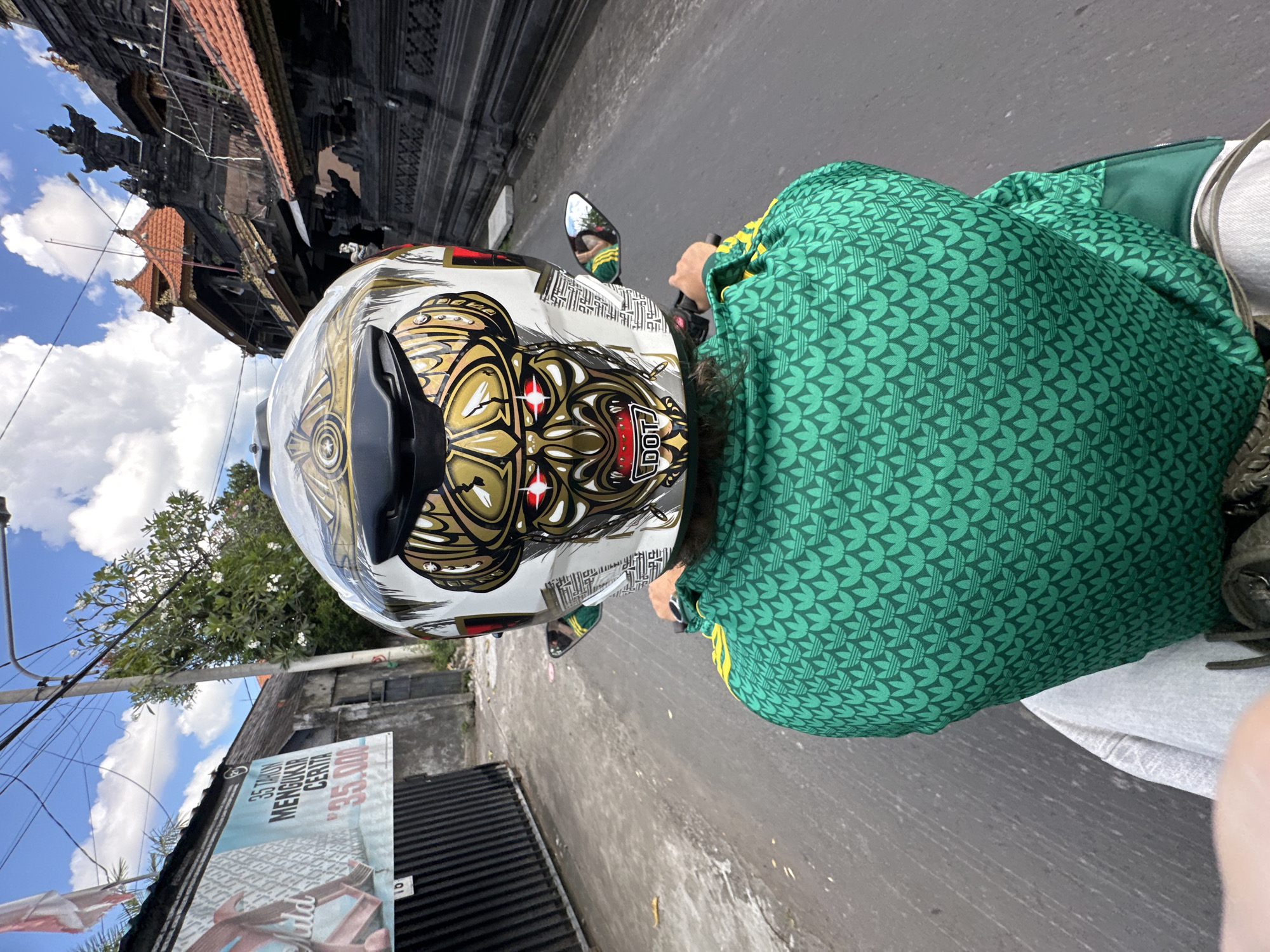 Captured from a high angle, the image shows a person riding a motorcycle along a gray asphalt road in North Kuta, Indonesia. The rider is wearing a green jacket with a repeating leaf-like pattern and a white helmet adorned with a gold and black graphic design that includes a face with prominent red eyes. A motorcycle mirror is visible on the left side of the helmet. In the background, buildings including one constructed of dark stone are juxtaposed against a blue sky scattered with white clouds. A large advertisement board is visible on the left side of the road near utility poles.