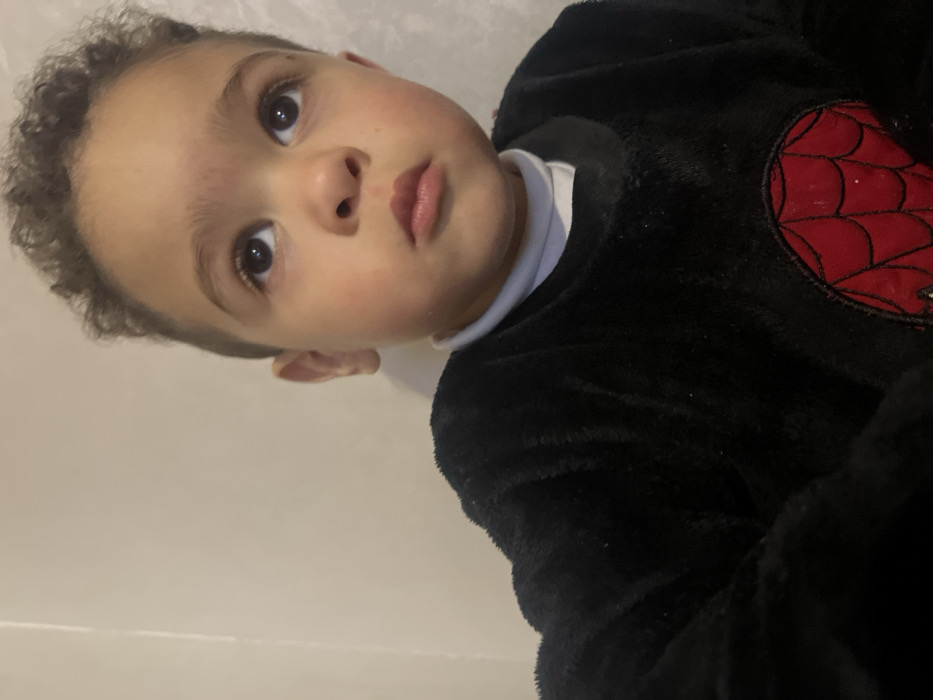 A young child with curly hair and brown eyes looks directly at the viewer. They have a fair complexion, a small nose, and slightly parted lips. The child is wearing a black, fluffy outfit with a white turtleneck underneath. A red spiderweb design is visible on the shoulder of the outfit. The background is a plain white or light grey wall, casting a soft light on the child's face.