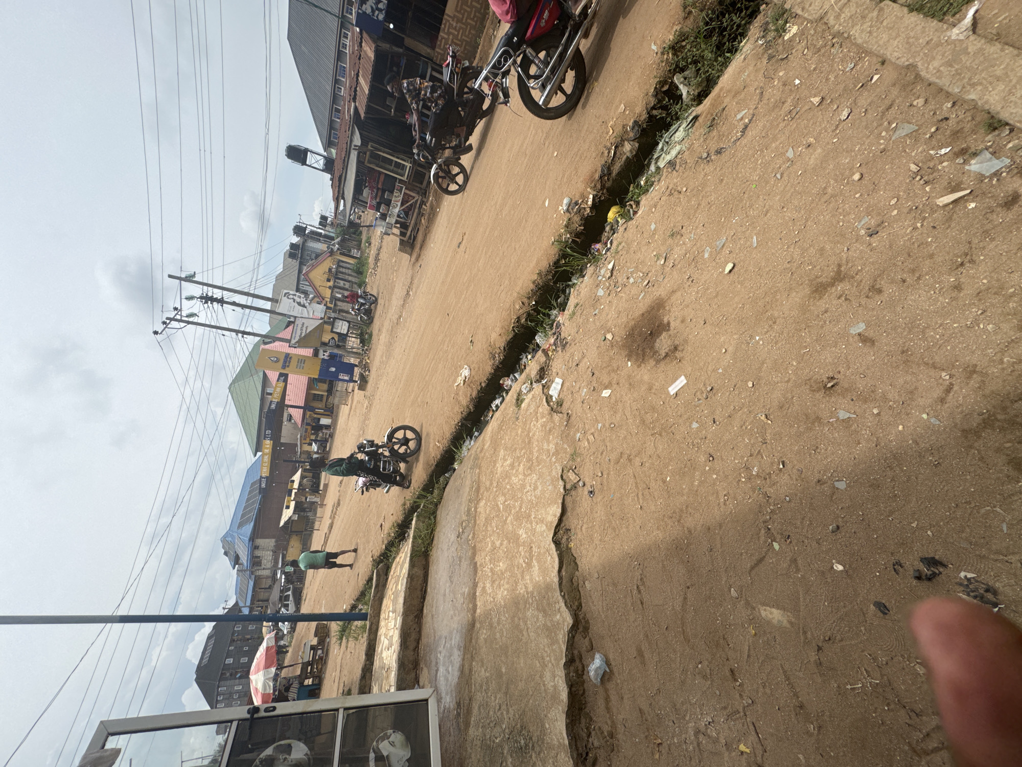 The image shows a street in Ihiagwa, Nigeria. The street is unpaved and dusty, with a deep drainage ditch running along the side. There is litter scattered along the street and in the ditch. Buildings line both sides of the street, and electrical wires are strung overhead. Several motorcycles are parked or moving along the street. A person is walking along the side of the road. Part of a window is visible in the foreground. The overall scene depicts a typical street scene in a developing urban area.