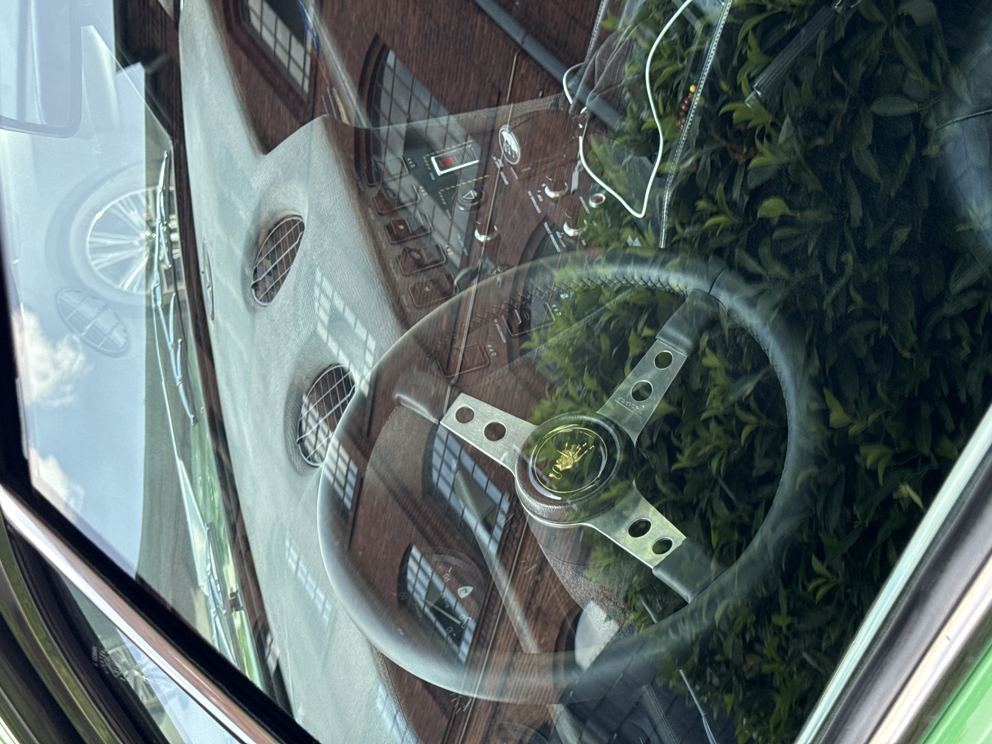 The image shows the interior of a vintage car, likely a classic sports car, viewed through the windshield. The steering wheel is prominent in the foreground, with a central hub featuring a golden Lamborghini emblem. The dashboard is visible, displaying various gauges and switches, also reflecting the brick building outside. A bright green bush is seen outside the car, partially obscuring the lower part of the image. The reflections in the windshield suggest the car is parked in a city environment with brick architecture. While the exact location within Offenbach am Main, Germany, is not determinable from the image alone, the context implies an urban setting. There are no people visible in the image.