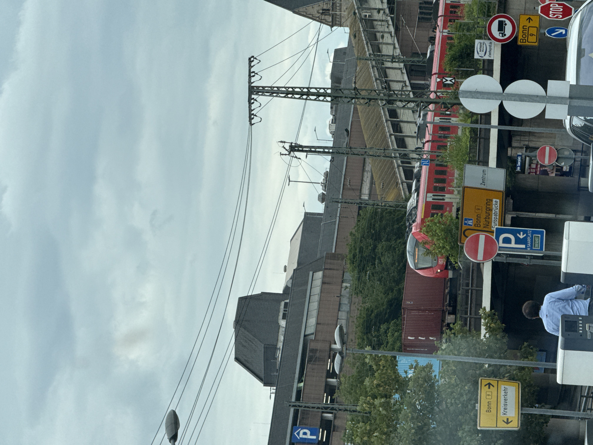 The image depicts a multi-level train station in Koblenz, Germany, under a cloudy sky. A red double-decker train is in motion on an elevated track. In the foreground, traffic signs indicate directions for Bonn and "Kreisverkehr" (roundabout), along with a parking sign. A person in a blue shirt is visible in the lower left of the frame, facing away from the camera. Power lines and electrical infrastructure are prominent throughout the scene, suggesting an urban environment. The architecture of the station appears to be modern concrete and glass, with some older brick structures visible in the background. The overall impression is one of an active transportation hub in a built-up area.