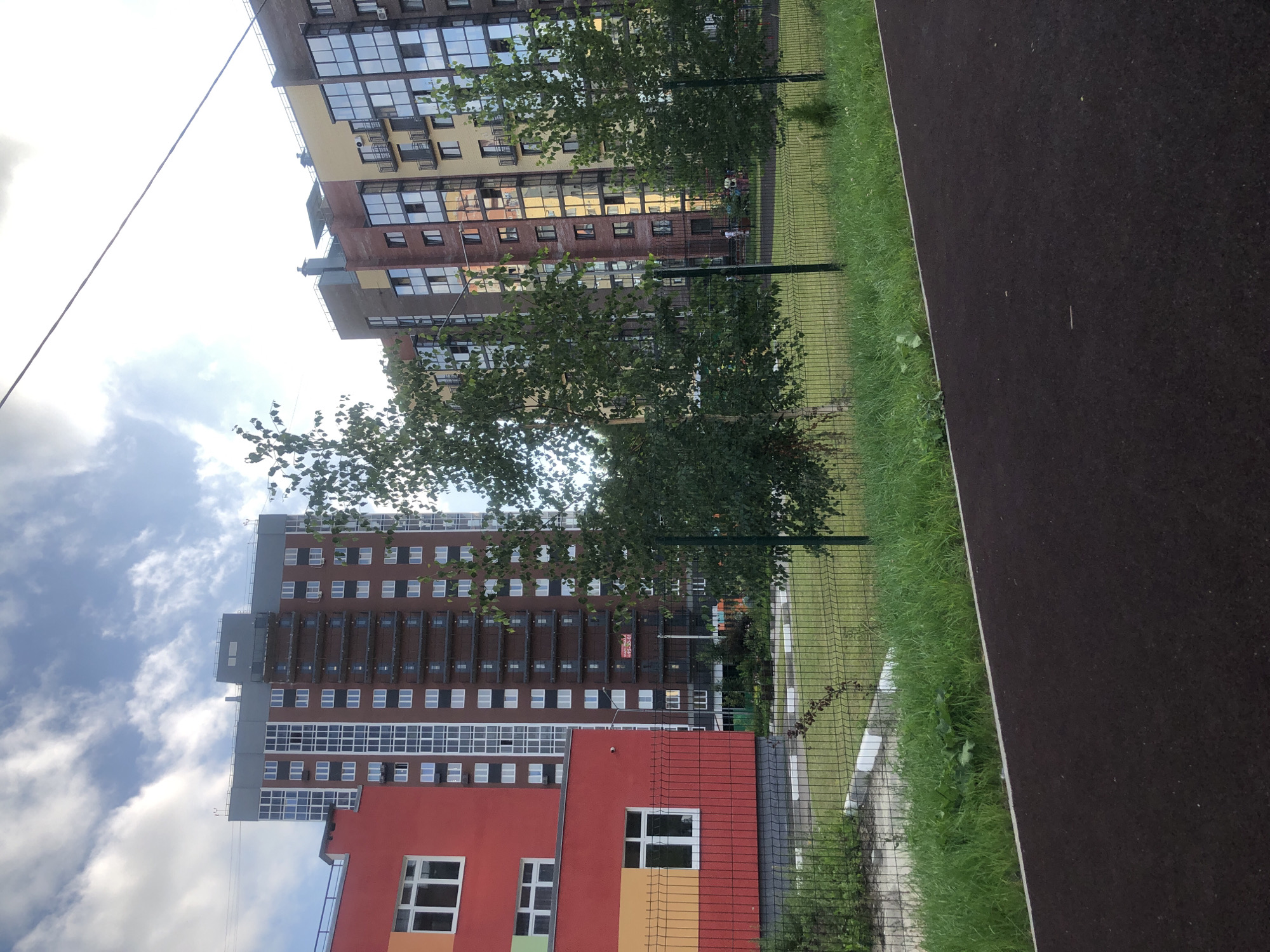 This is a photograph of a modern residential complex in Fedorino, Russia, under a cloudy sky. In the foreground, a dark asphalt surface, possibly a running track or playground area, is visible. A metal mesh fence separates this area from a green lawn and a paved walkway with white bollards. Beyond the fence, two buildings dominate the scene. To the left is a low-rise building with a red and yellow facade, featuring white-framed windows. To its right stands a tall, multi-story apartment building with a reddish-brown brick exterior and numerous white-framed windows. Further to the right is another apartment building with a colorful facade, featuring yellow, brown, and brick sections with large glass windows and balconies. Several trees, including birch trees, are interspersed throughout the scene, with their branches reaching towards the buildings. In the background, faint outlines of other structures and utility poles are visible.