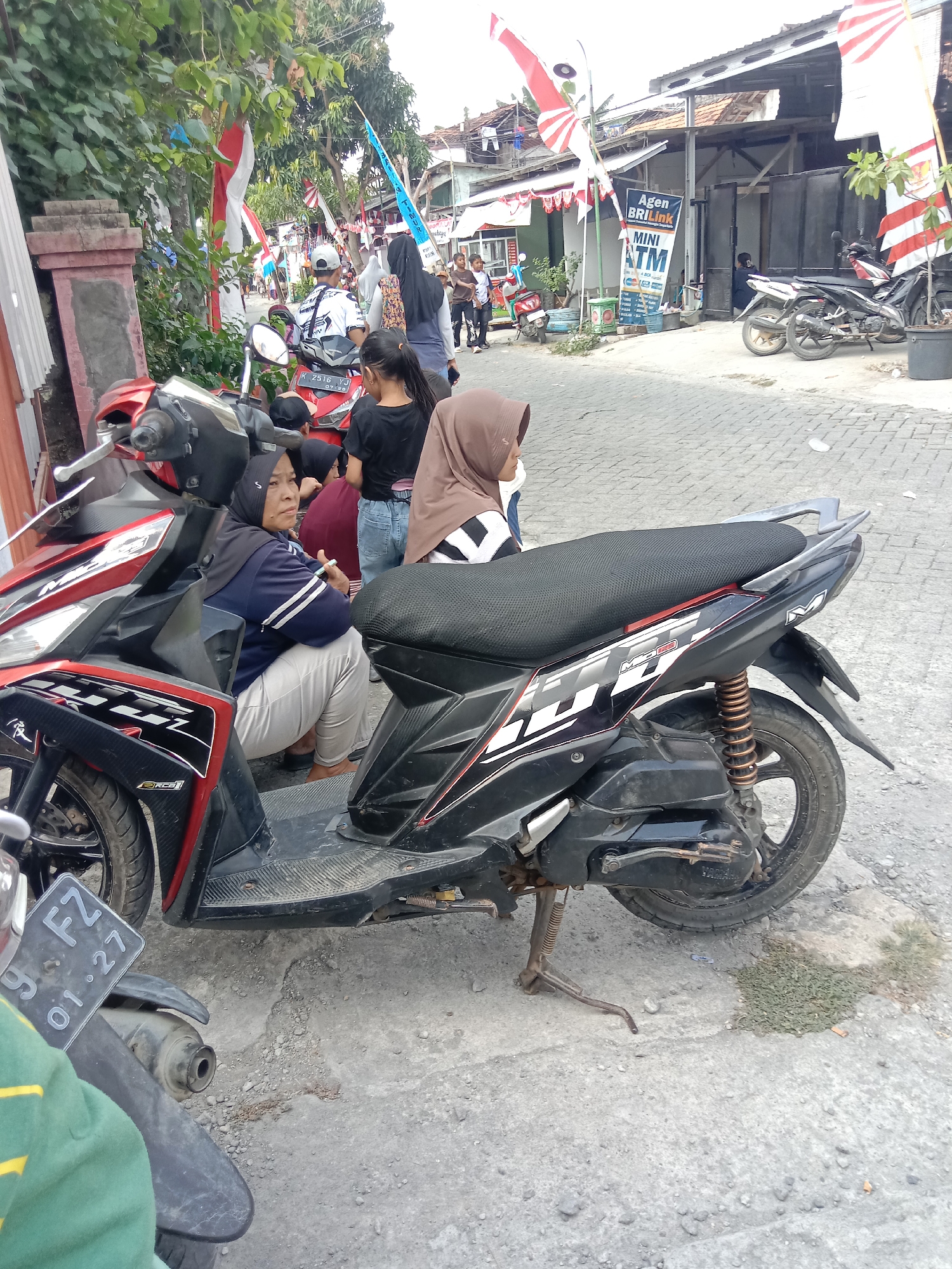 A street scene in Wirosari, Indonesia, features several people and motorcycles. In the foreground, a red and black motorcycle is parked, with a woman wearing a blue jacket and light-colored pants seated on it. To her right, another motorcycle, predominantly black, is also parked. Behind these motorcycles, several other people are visible, including a woman in a brown hijab and a person wearing a white t-shirt and a cap. In the background, buildings line the street, adorned with Indonesian flags and banners, suggesting a local celebration or event. A sign for "Agen BRI Link MINI ATM" is prominently displayed on one of the buildings. The street is paved with interlocking concrete blocks.