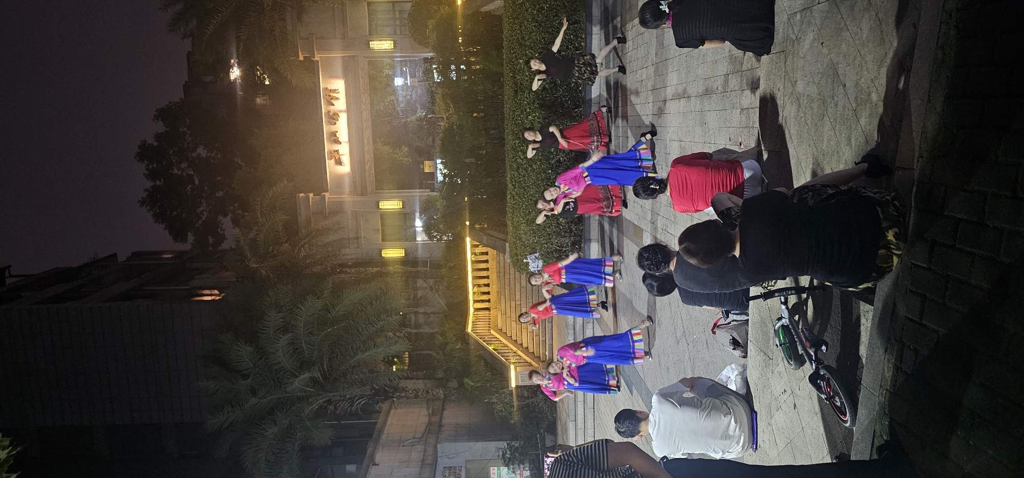 This image depicts an outdoor nighttime scene in Jiulongpo District, China, featuring a group of people dancing and an audience watching. The dancers, all women, are dressed in colorful traditional attire, predominantly bright pink tops and long blue skirts with decorative borders. They are positioned on a paved plaza in front of a lush green hedge and a building with ornate architecture, illuminated by yellow lanterns and a large sign with Chinese characters. The dancers appear to be performing a choreographed routine, with several in mid-motion.
In the foreground, seated on the ground, are several spectators. They are facing the dancers, some with their backs to the camera. These spectators are casually dressed. To the left, a person is partially visible, holding a phone, suggesting they are recording or photographing the performance. A child's scooter is lying on its side on the plaza near the seated spectators. The environment is characterized by palm trees and urban architecture, with artificial lighting providing illumination for the performance and the surrounding area. The sky is dark, indicating it is nighttime.