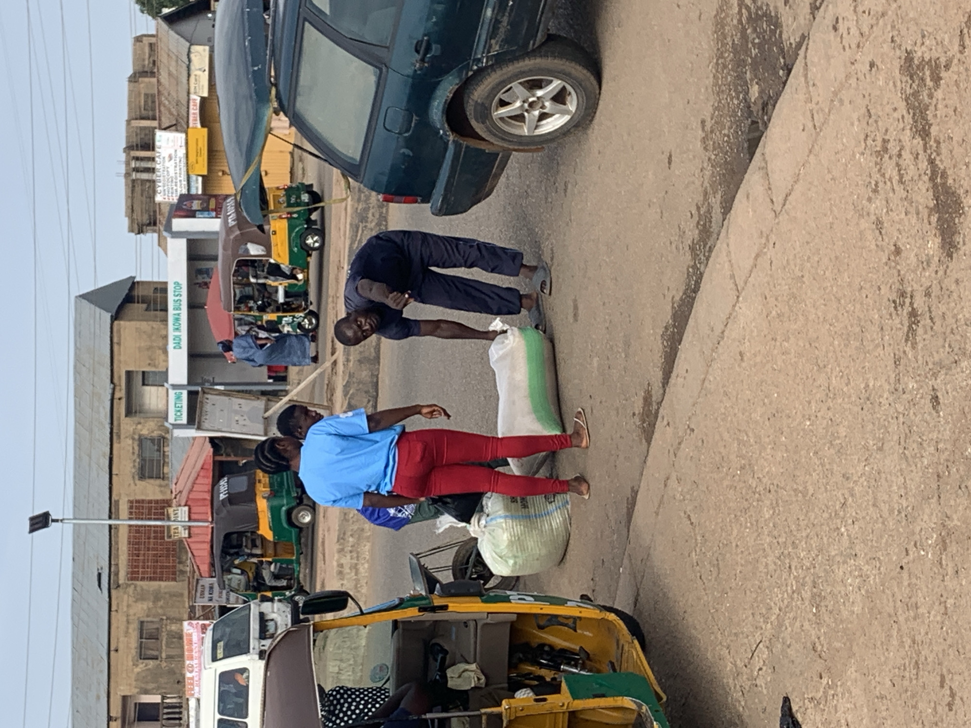 In Gura Top, Nigeria, an outdoor scene unfolds at what appears to be a bus stop with a "DADI IKOWA BUS STOP" sign. A man in a dark blue t-shirt and pants is actively engaged in lifting a large, light-colored sack, pointing forward with his right hand. Beside him, a person in a blue t-shirt and red pants stands with their back to the camera, looking towards the sack. Another individual in a green and yellow auto-rickshaw, also known as a "Keke Napep," is partially visible, with a person in traditional blue attire standing near its open door. To the right, the rear of a dark green station wagon is parked on the asphalt road, its driver's side rear wheel clearly visible. The environment features a concrete sidewalk, road surface with visible patches and cracks, and buildings in the background with signs that include "FEEL GO HOME," "TEA DINT," and "CYBER CAFE." A lamppost with a solar panel is also present.