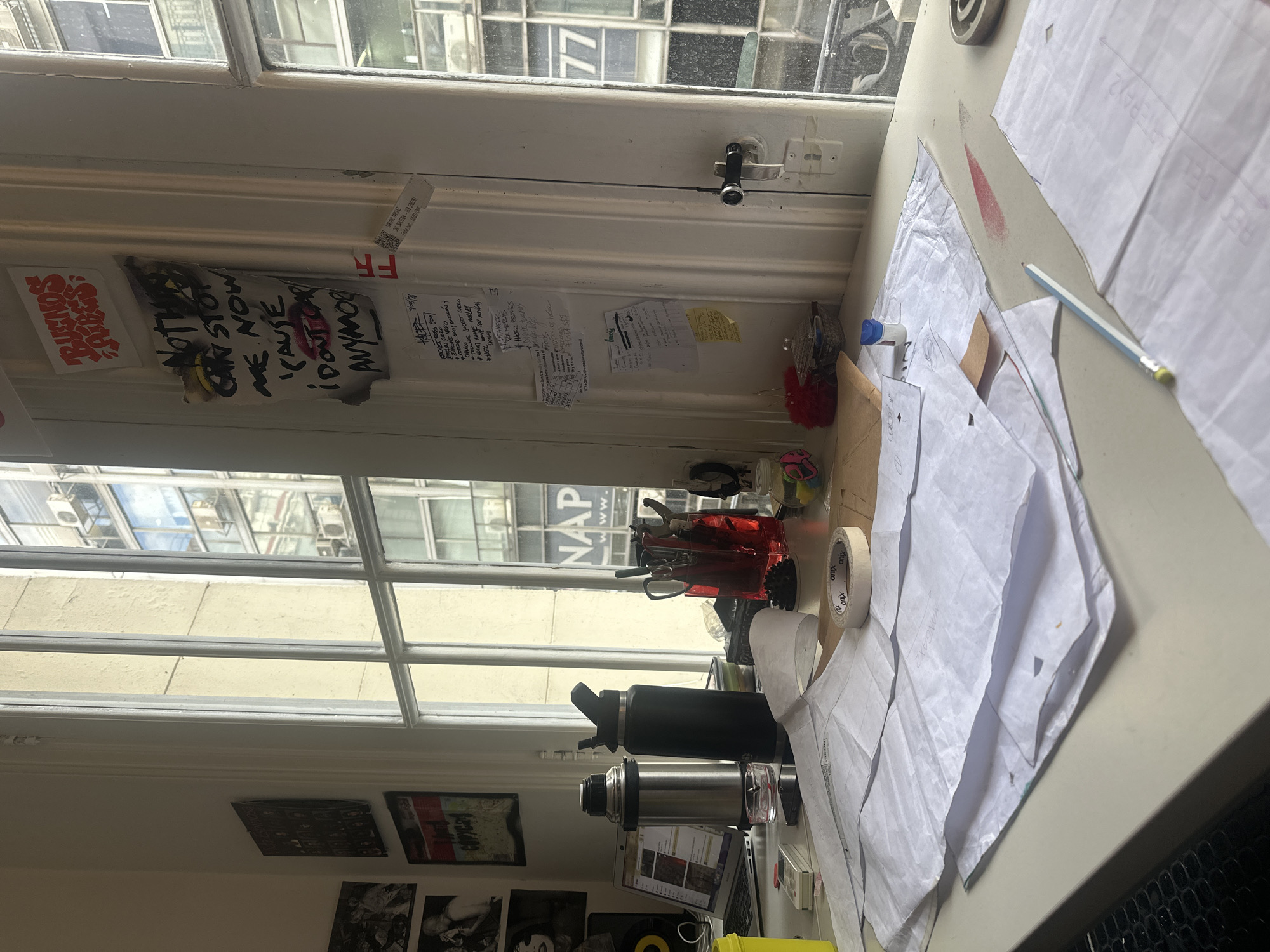 The image shows a cluttered workspace next to a window. Several sheets of white paper, possibly patterns, are spread across the surface. A pencil lies on top of the papers. Nearby are rolls of tape, a red pen container with scissors and pens, a black water bottle, a silver thermos, and a laptop. The window looks out onto a street in a city, with a building visible across the street displaying the word "NAP." On the window frame are stickers and notes, including a sticker with "BUENOS AIRES" written on it. Below the window are framed images or posters on the wall. Based on the sticker indicating "BUENOS AIRES," it is likely this workspace is located in Buenos Aires, Argentina.
