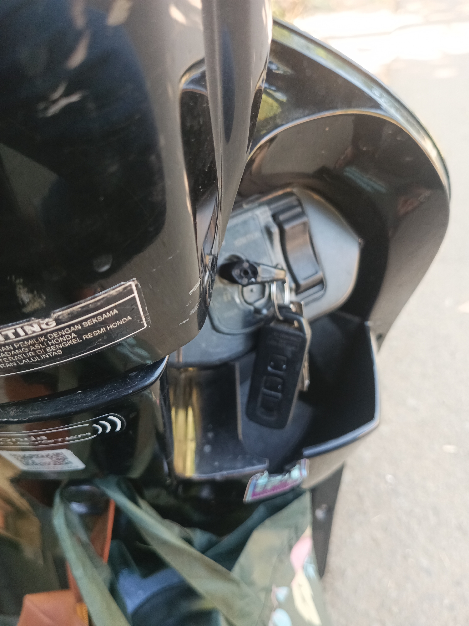 In Tumampua, Indonesia, a close-up shot focuses on the ignition and storage compartment of a black motorcycle. The key is inserted into the ignition, with a second key attached to a small black fob hanging from it. The storage compartment below appears empty, revealing the inner plastic lining of the motorcycle. A portion of a white sticker with text in Indonesian is visible on the left side of the image, and some scuff marks are present on the black plastic body of the motorcycle. The background is out of focus, showing a neutral, outdoor setting, likely a street.