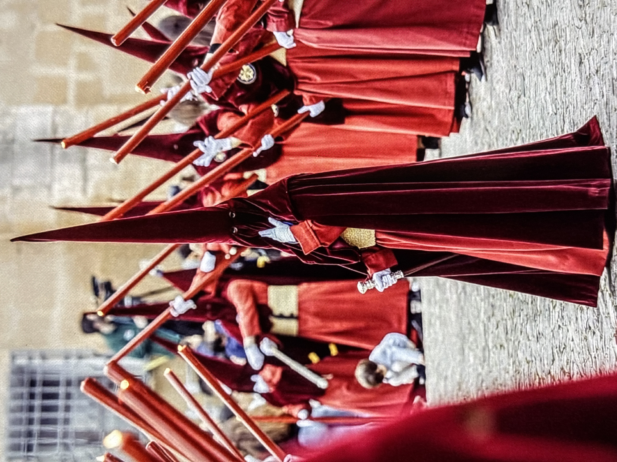 The image depicts a religious procession in the city of Orihuela, Spain. Several individuals, dressed in traditional red hooded robes and capes, are participating. They are holding tall red poles, some of which appear to be lit. The ground is paved with cobblestones, indicative of a historic urban setting. In the background, a person is visible operating a video camera, suggesting the event is being documented. A young child, dressed in light blue, is seen crouching on the ground amongst the procession participants. The architecture in the far background features windows with grates and stone walls. The overall atmosphere suggests a formal and solemn religious observance.