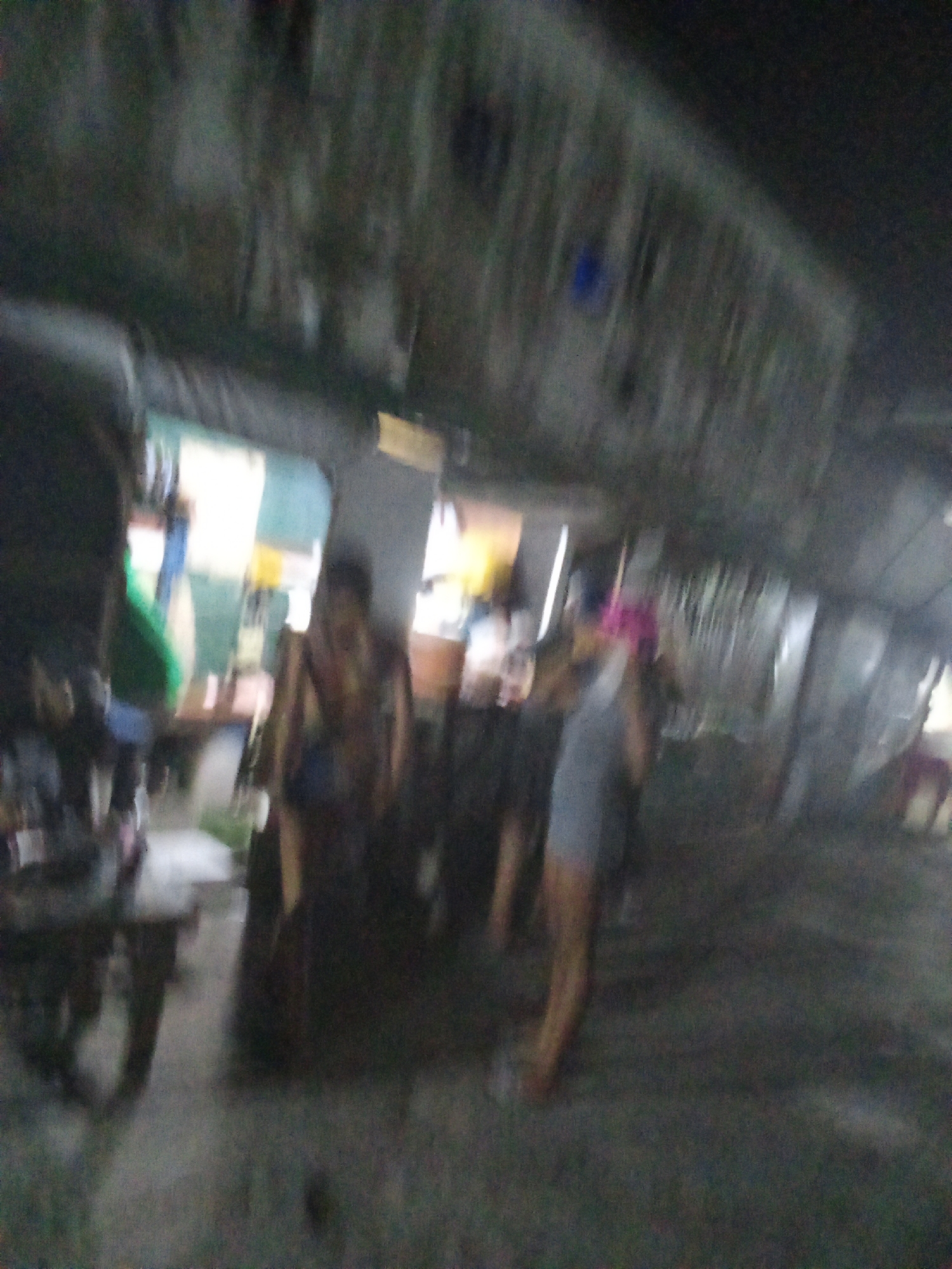 The image, likely taken in Ado, Nigeria, shows a blurry street scene at night. Two figures are discernible: one wearing a dark tank top, and another in a gray outfit with a pink head covering. The setting is an outdoor space, possibly a market stall or a shopfront, with tables, chairs, and other indistinct objects scattered around. The area appears dimly lit, suggesting it may be an underserved part of the city.