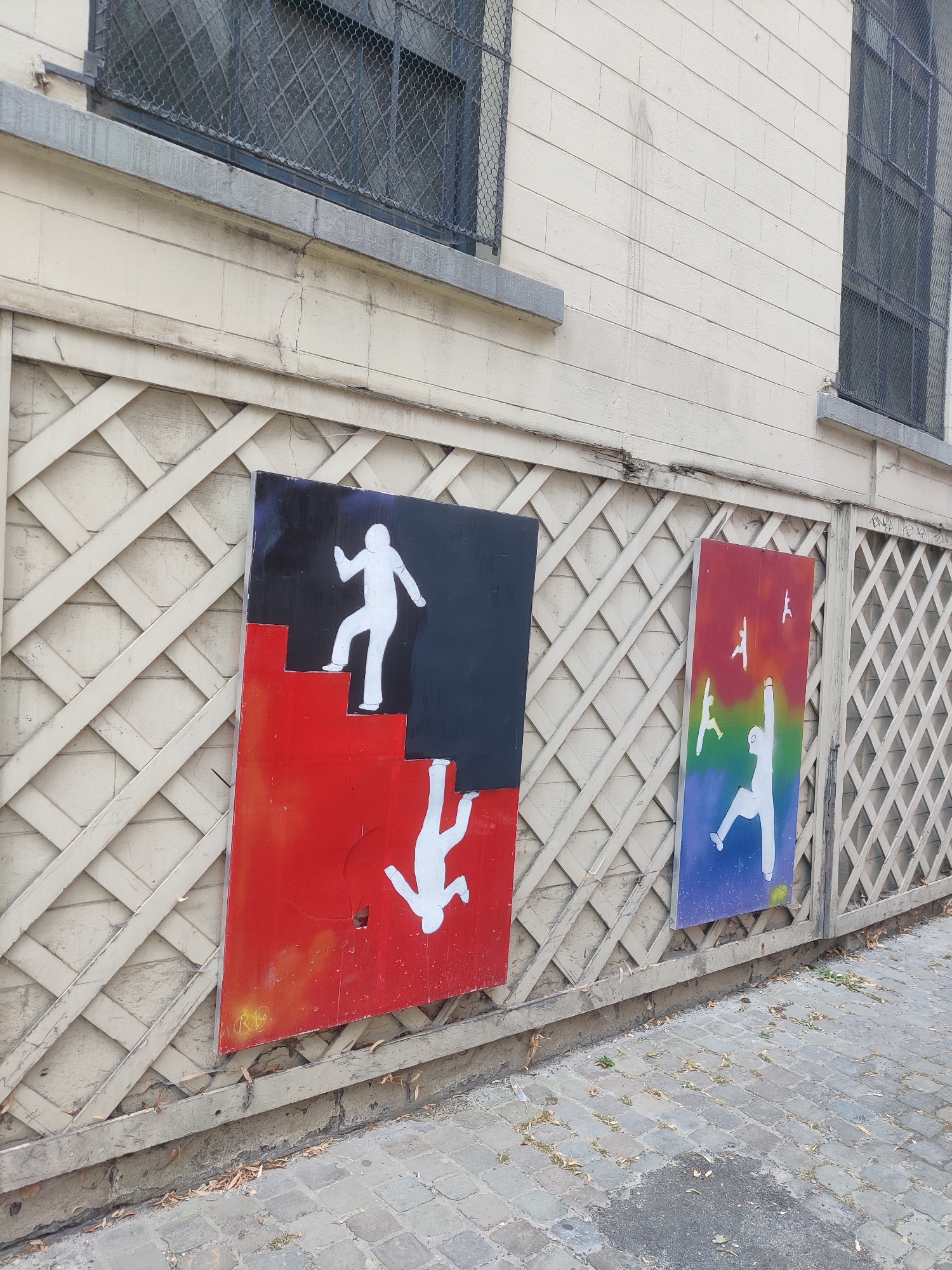 The image shows two artworks mounted on a beige wooden lattice fence. The artwork on the left is a large rectangular panel divided into two sections: a black upper section and a red lower section. A white silhouette of a person is depicted on stairs, ascending from the red section into the black section. Below the stairs, another white silhouette is shown falling. The artwork on the right is a rectangular panel with a rainbow gradient background. Several white silhouettes of people are depicted in various dynamic poses, some appearing to be falling or leaping. The fence is attached to a building with light-colored horizontal siding and barred windows. The ground in front of the fence is paved with cobblestones. The scene appears to be an outdoor urban environment. Based on the provided information, this location is in the city of Liège, Belgium.