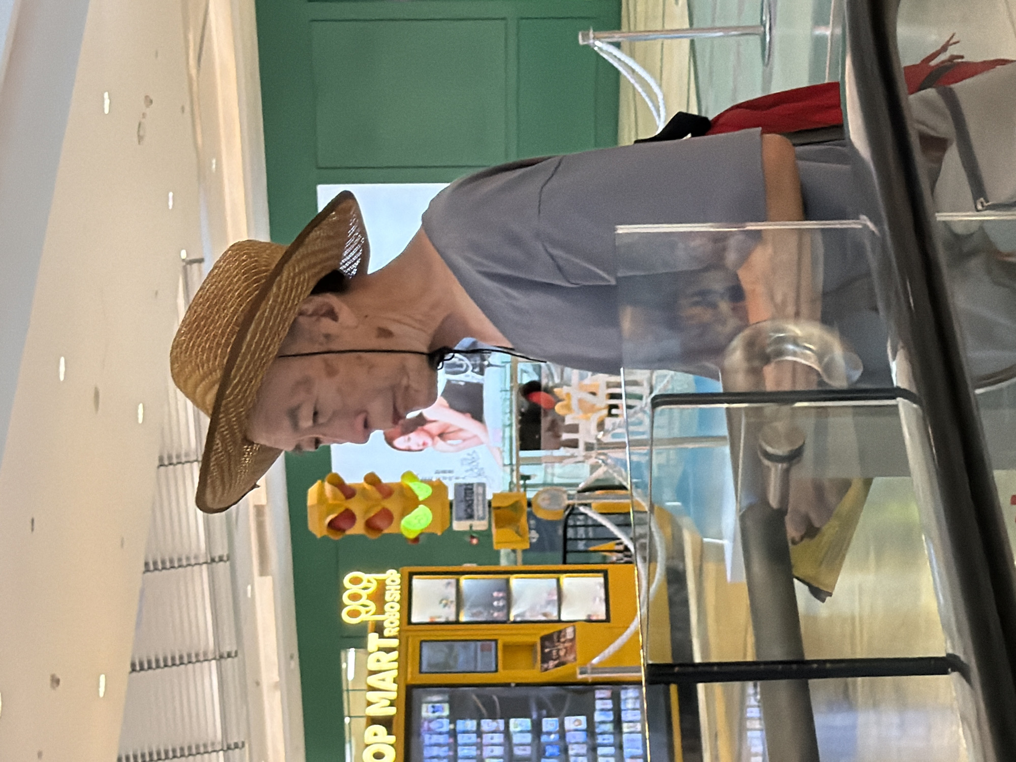 An elderly East Asian man wearing a straw hat and a grey t-shirt is standing in front of a yellow "POP MART ROBO SHOP" vending machine, possibly in a shopping mall in Yuzhong District, China. He is looking to his right. The vending machine displays various toys. A traffic light-shaped decoration is also visible to the right of the vending machine. The man appears to have some discoloration on his face and neck. He is partially obscured by a glass barrier. In the background, there are green walls and a large advertisement featuring a woman. The overall environment suggests an indoor commercial setting.