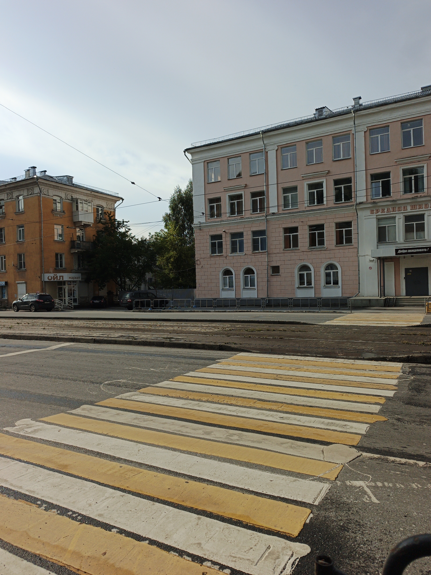 The image shows a street scene in Zlatoust, Russia. In the foreground, a yellow and white striped pedestrian crossing is visible on an asphalt road. Beyond the crossing, tram tracks run parallel to the road.
To the left of the tram tracks is a multi-story orange building with multiple windows and balconies. A sign on this building reads "ОЙЛ маркет" (OIL market). A dark-colored car is parked in front of the building.
To the right of the orange building, and dominating the middle and background of the image, is a large, light pink multi-story building with many windows. This building appears to be a school, as suggested by signage which includes "СРЕДНЯЯ ШКОЛА" (Secondary School) and "Добро пожаловать!" (Welcome!). A dark doorway is visible on the ground floor of this building.
Several trees are visible behind the orange building and between the two main buildings. The sky is bright and appears to be partly cloudy. The overall impression is of an urban environment on a clear day.