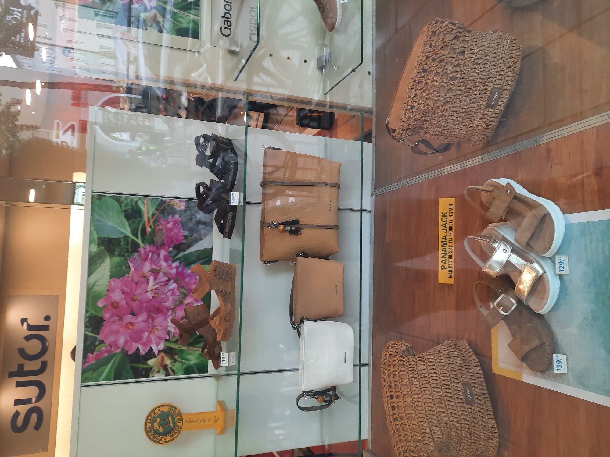 The image depicts a storefront display, likely a shoe or fashion boutique, featuring various items for sale. The "Sutor" logo is prominently displayed at the top of the window. On the first shelf, there are two pairs of women's sandals, one tan wedge sandal with cork soles priced at "119.99", and a black wedge sandal with a similar sole. A circular "Panama Jack, Made in Spain" emblem is visible to the left.
The second shelf holds three handbags. From left to right, there's a small white crossbody bag, a medium-sized tan leather handbag with decorative charms, and another tan handbag that appears to be made of a textured material.
The bottom portion of the display showcases two pairs of women's sandals. One is a tan sandal with a buckle closure and cork sole, priced at "139.99". The other is a metallic gold sandal with a white platform sole, priced at "129.99". To the left of these sandals is a woven straw tote bag. Another woven straw crossbody bag is visible to the right, with a logo that reads "Panama Jack". A yellow sign with the text "PANAMA JACK MANUFACTURES ALL ITS PRODUCTS IN SPAIN" is positioned between the woven bags and the sandals.
The overall environment is that of a retail display, with clear glass shelves and a wooden floor. The reflection in the glass suggests an outdoor setting, possibly a street with buildings and trees, indicating the location is likely in a city or town. Considering the presence of the "Sutor" brand, which has multiple locations, and the general architectural style visible in the reflections, the store is located in Rosenheim, Germany.