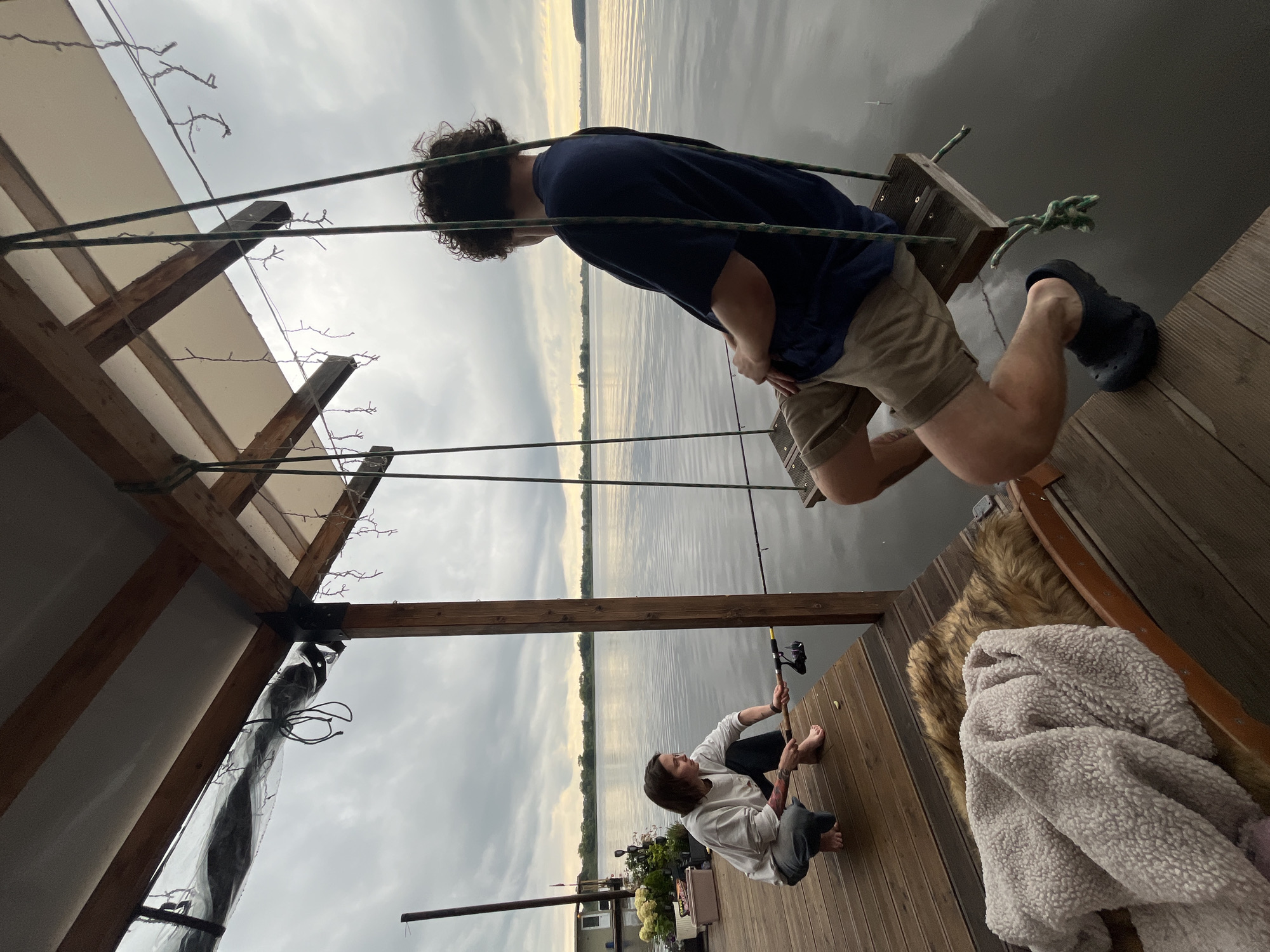 On a wooden dock on a cloudy day, two people are present. One individual, with short brown hair and tattoos on their left arm, is squatting on the dock, holding a fishing rod. They are wearing a light grey sweatshirt and dark pants. To their right, another person with curly brown hair is sitting on a swing. This individual is wearing a navy blue t-shirt and khaki shorts. The swing is suspended from a wooden structure above, which has some string lights attached. The dock extends over a body of water, with a calm surface reflecting the overcast sky. In the background, a floating house is visible on the left side of the image, and a treeline can be seen in the distance. The overall scene appears to be in a natural, waterside environment, likely a rural area or a location outside of a major city. The mention of Radchenko, Russia is not supported by visual evidence in the image.