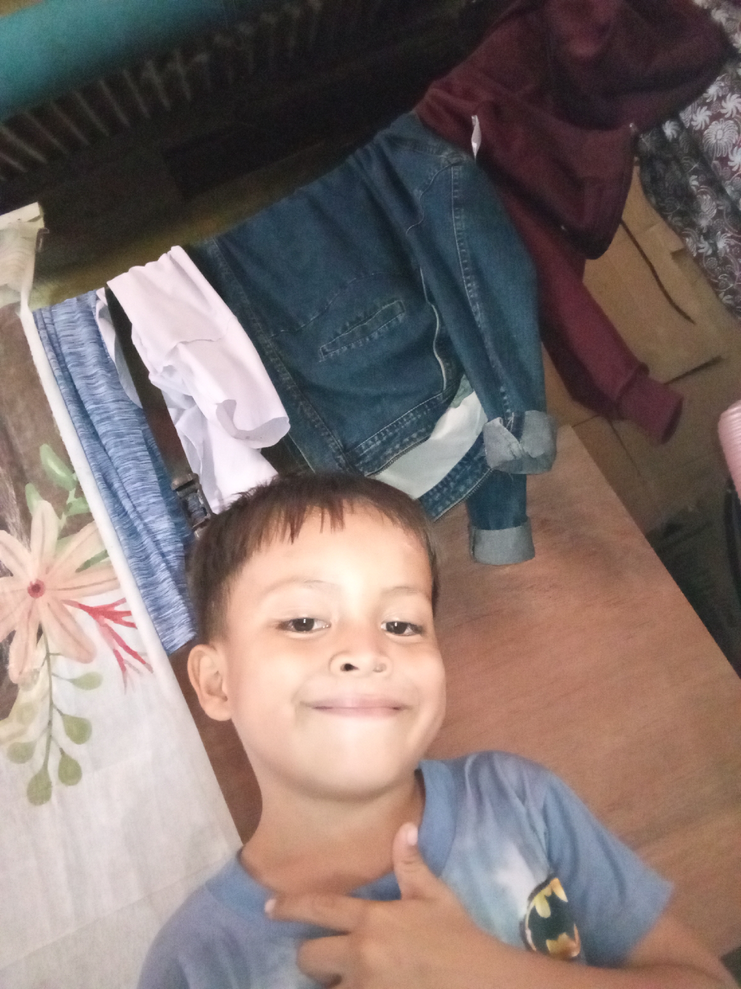 A young boy with short, dark hair and a light complexion is pictured in a close-up shot, smiling and pointing at his chest. He is wearing a light blue t-shirt with a Batman logo on it. The background consists of clothing hanging on a clothesline, including a denim jacket and a maroon hoodie. A wooden surface, possibly a table or floor, is visible below the boy and the hanging clothes. The overall impression is that of an indoor scene, likely a domestic setting.