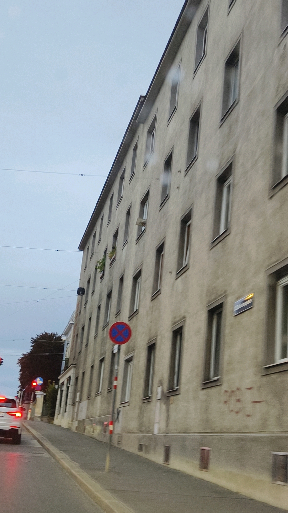 The image shows a street scene in Vienna, Austria, with a tall, pale apartment building on the right side of the frame. The building has a facade with numerous windows, many of which are open or have shutters. A traffic light is visible on the left, showing red. A white car is stopped at the intersection, its brake lights illuminated. Several street signs are present, including a "no parking" sign. The sky is overcast, suggesting a cloudy day. The building appears to be several stories high and has a uniform architectural style. There is some graffiti visible on the lower part of the building's facade. The street is paved and there is a sidewalk.