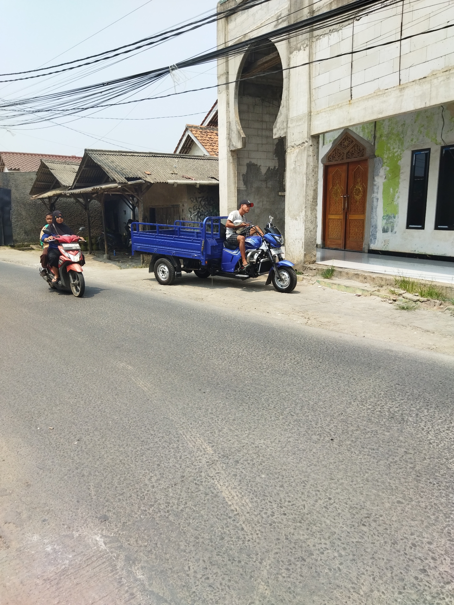 In Suka Sari, Indonesia, a bright blue three-wheeled vehicle with a cargo bed is parked on the right side of a paved road. A man wearing a white t-shirt, dark shorts, and a red baseball cap is seated on the driver's side of the vehicle, holding what appears to be a mobile phone. Further down the road on the left, a red scooter is being ridden by a woman wearing a dark hijab and a patterned shirt, with a child seated behind her. The background features several buildings with corrugated metal roofs, and a large building with a prominent arched entrance and carved wooden doors. Overhead, a dense network of electrical wires stretches across the frame. The sky is clear and bright, indicating daytime.