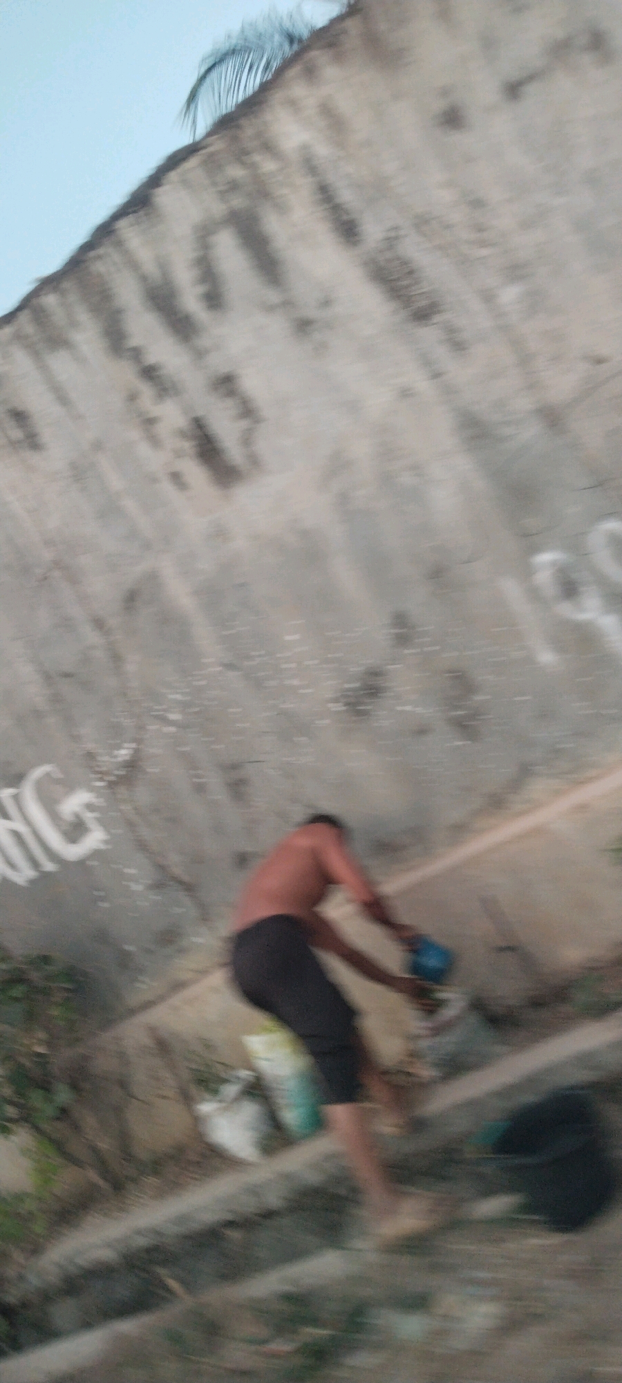 A person is captured in motion, appearing to be shirtless and wearing dark pants, in the vicinity of Tanjung Ratu Ilir, Indonesia. The individual is interacting with a blue object, possibly a tool or container, near a concrete structure that shows signs of graffiti. The background consists of a textured concrete wall, with some vegetation visible at the lower left and right. The image is blurry, suggesting movement or a low-quality camera.