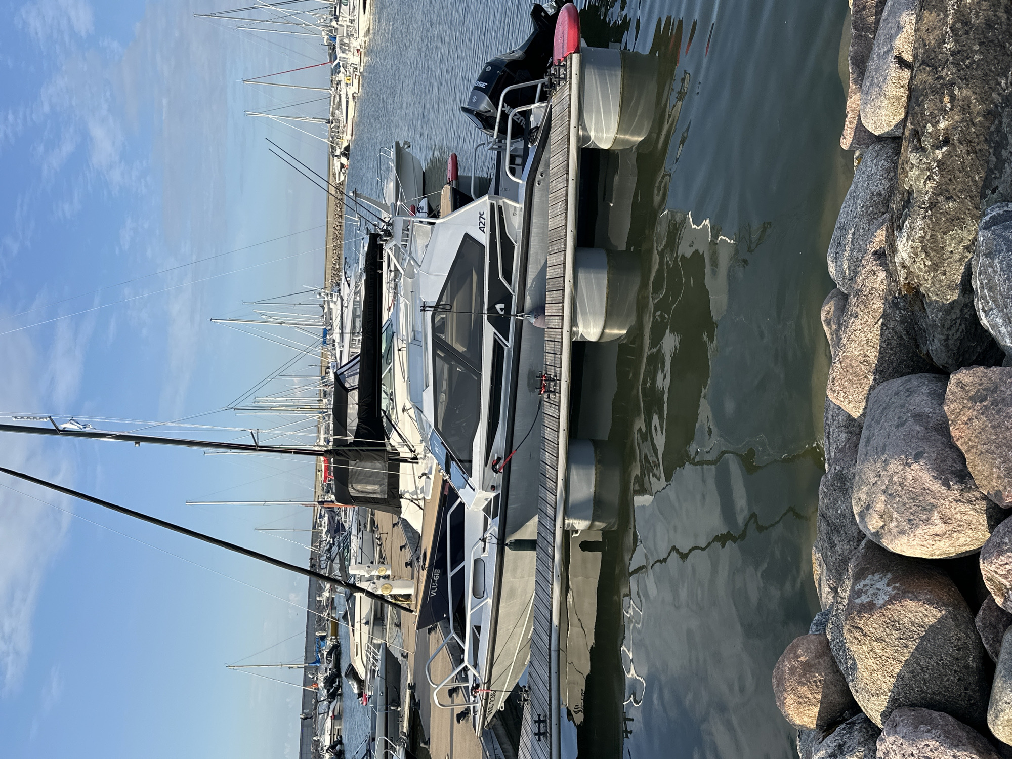This is a medium shot of a marina on a clear day, likely in Tallinn, Estonia, given the flag. In the foreground, a modern motorboat with a black and white cabin is docked. The boat is gray with black accents and has a Suzuki outboard motor. It is secured to a wooden dock with floating pontoon supports. Next to it, another gray boat with the registration VLU-618 is visible. The water is calm and reflects the sky and the boats. The background is filled with numerous sailboats and other motorboats moored in the marina. A stone breakwater stretches across the far background, and a clear blue sky with some wispy clouds is overhead. Large, rough-hewn rocks are piled along the edge of the water in the immediate foreground. There are no people clearly visible in the image. The overall scene suggests a peaceful and well-maintained maritime setting.