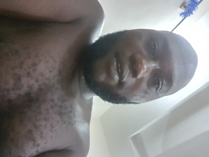 An adult dark-skinned male is depicted in a close-up, upward-angled shot. He has a bald or very closely shaven head and a full dark beard and mustache. He is bare-chested, displaying scattered dark hair on his chest and shoulders. A small drop of liquid is visible on his beard near his Adam's apple. His eyes are directed towards the camera, and his mouth is slightly ajar. The background consists of a light-colored wall and ceiling. A blue, fluffy decorative item is affixed to the upper left wall. The location is an indoor setting within the city of Unknown, Unknown.
