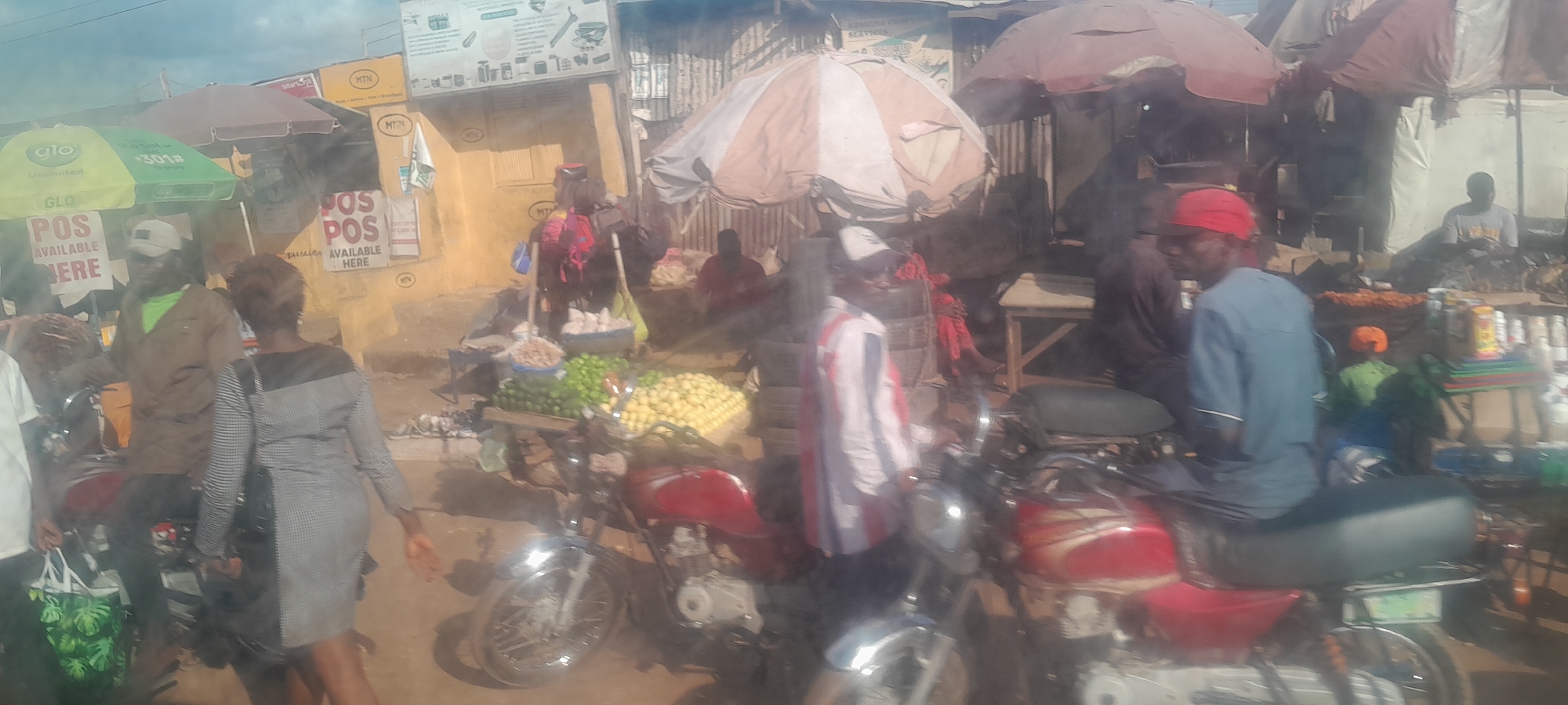 The image depicts a bustling market scene in Nyanya, Nigeria. In the foreground, two red motorcycles are parked side-by-side. The motorcycle on the right is more visible, with a black seat and visible chrome accents. To the left of the motorcycles, a vendor is selling produce, with piles of green items, possibly limes or cucumbers, and yellow items, likely lemons or yams, displayed on a wooden table. Behind the produce, a person in a grey and black dress is walking away from the camera, carrying a black bag. To the left of this person, a man in a brown jacket and a white cap is visible, seemingly interacting with someone off-camera. Further back, under a striped umbrella, a stall is set up with various items, and more people are gathered. Signs for "GLO" and "POS AVAILABLE HERE" are visible on the left side of the image. The background shows buildings with corrugated iron roofs and yellow walls, indicative of a market in a developing urban area. The overall impression is a lively and active marketplace.