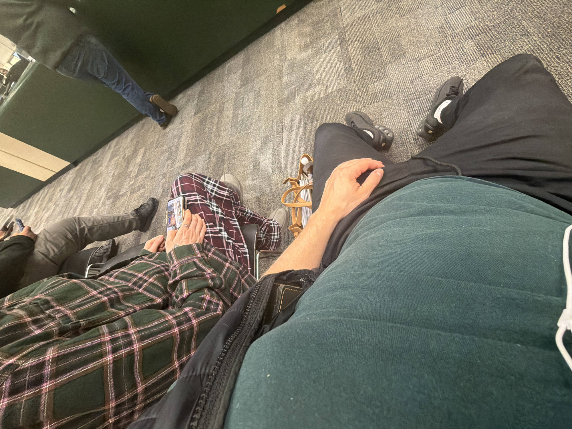 The image, taken from a first-person perspective, shows an interior scene of people sitting. In the foreground, a person wearing black pants and a dark green shirt is visible. To their left, another individual is wearing a green and pink plaid shirt and maroon plaid pants. The legs of other people are visible further in the background, some wearing jeans and others wearing gray pants. The setting appears to be an airport, specifically O'Hare International Airport near the city of Skokie, United States of America, possibly a waiting area or terminal due to the presence of seated individuals and luggage near them. The floor is covered in a light gray carpet.