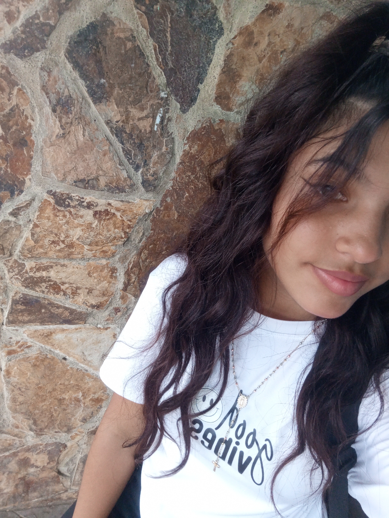 A young woman stands against a stone wall in Altagracia, Venezuela. The wall is composed of irregularly shaped stones of various brown and tan hues, mortared together. The woman has long, dark, wavy hair and is wearing a white t-shirt with a printed design and a silver necklace featuring a small pendant. Part of a black backpack strap is visible over her right shoulder. Her expression is neutral, and she is looking slightly off-camera.