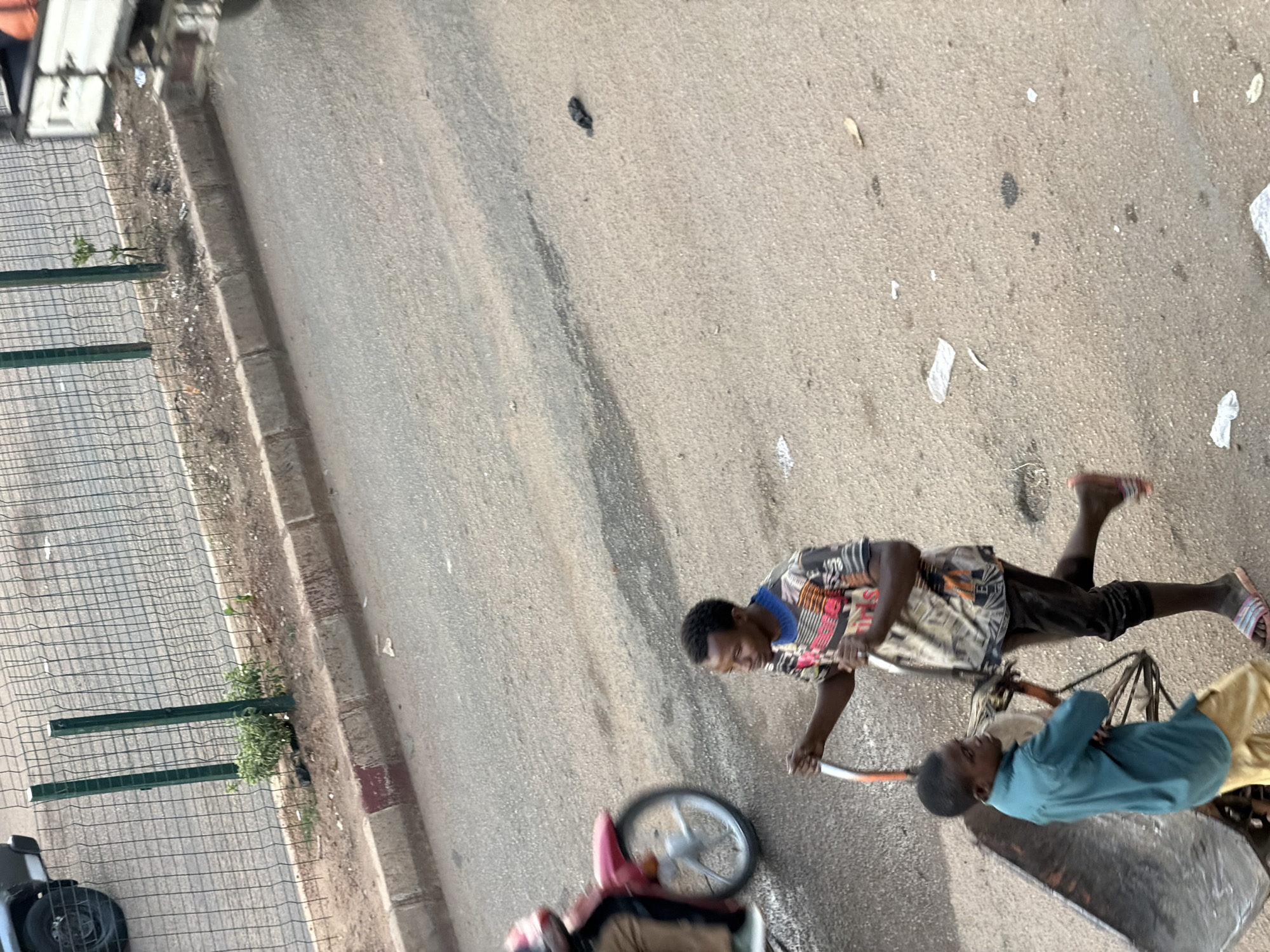The image shows two young boys in Bukuru, Nigeria, appearing to be moving a wheelbarrow. One boy, wearing a colorful patterned shirt and dark shorts, is pushing the wheelbarrow. The other boy, in a blue shirt and dark shorts, is sitting in the wheelbarrow. The scene is an outdoor street with an asphalt surface and a sidewalk. A wire fence with some green vegetation growing through it is visible on the left side of the image. Debris and scattered white paper or plastic are present on the ground. The lighting suggests it is daytime, likely sunny. The expression on the boy pushing the wheelbarrow seems focused, while the boy sitting appears relaxed. No visible text is present in the image.