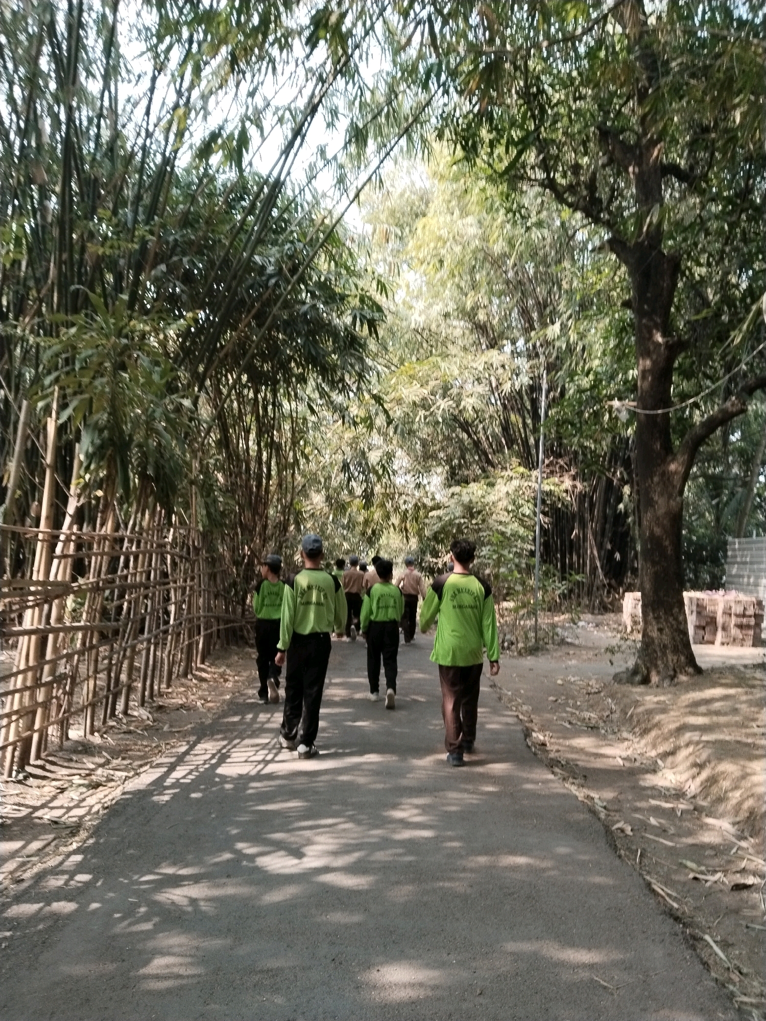 A group of seven young individuals wearing matching light green shirts and dark pants are walking away from the camera down a paved path. The path is flanked by dense bamboo on the left, with a woven bamboo fence running alongside it. On the right, there is a large tree and some bricks stacked, suggesting construction or material storage. The scene is outdoors, likely in a rural or park-like setting with dappled sunlight filtering through the trees and bamboo. The location is identified as Pakulaut, Indonesia.
