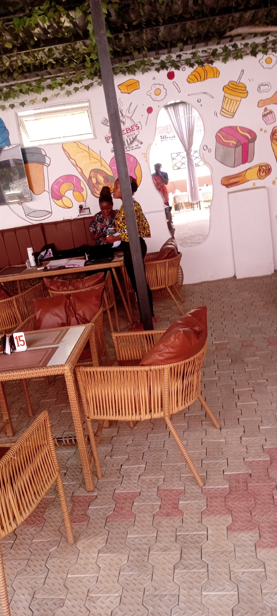 Two women are seated at a table in an outdoor seating area of a restaurant in Sabon Gari East, Nigeria. The restaurant wall has cartoon images of food, and there is a table marker with the number "15" visible. Chairs are made of woven material with leather cushions. The floor is paved with interlocking concrete blocks. A person is visible in a mirror on the wall. One woman is standing in the process of speaking.