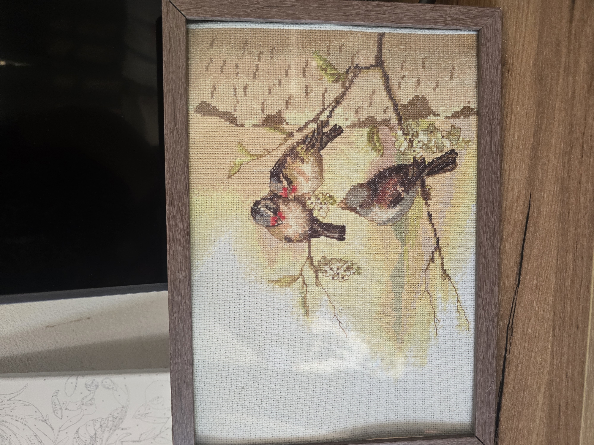 A framed cross-stitch embroidery of three birds on a branch is displayed. The birds, rendered in detail with brown, gray, white, and red coloration, are perched on a tree branch adorned with small white blossoms. The background depicts a soft, diffused light, suggesting a natural outdoor setting. The embroidery is housed in a simple brown frame and is positioned on a wooden surface, likely a table or shelf.
