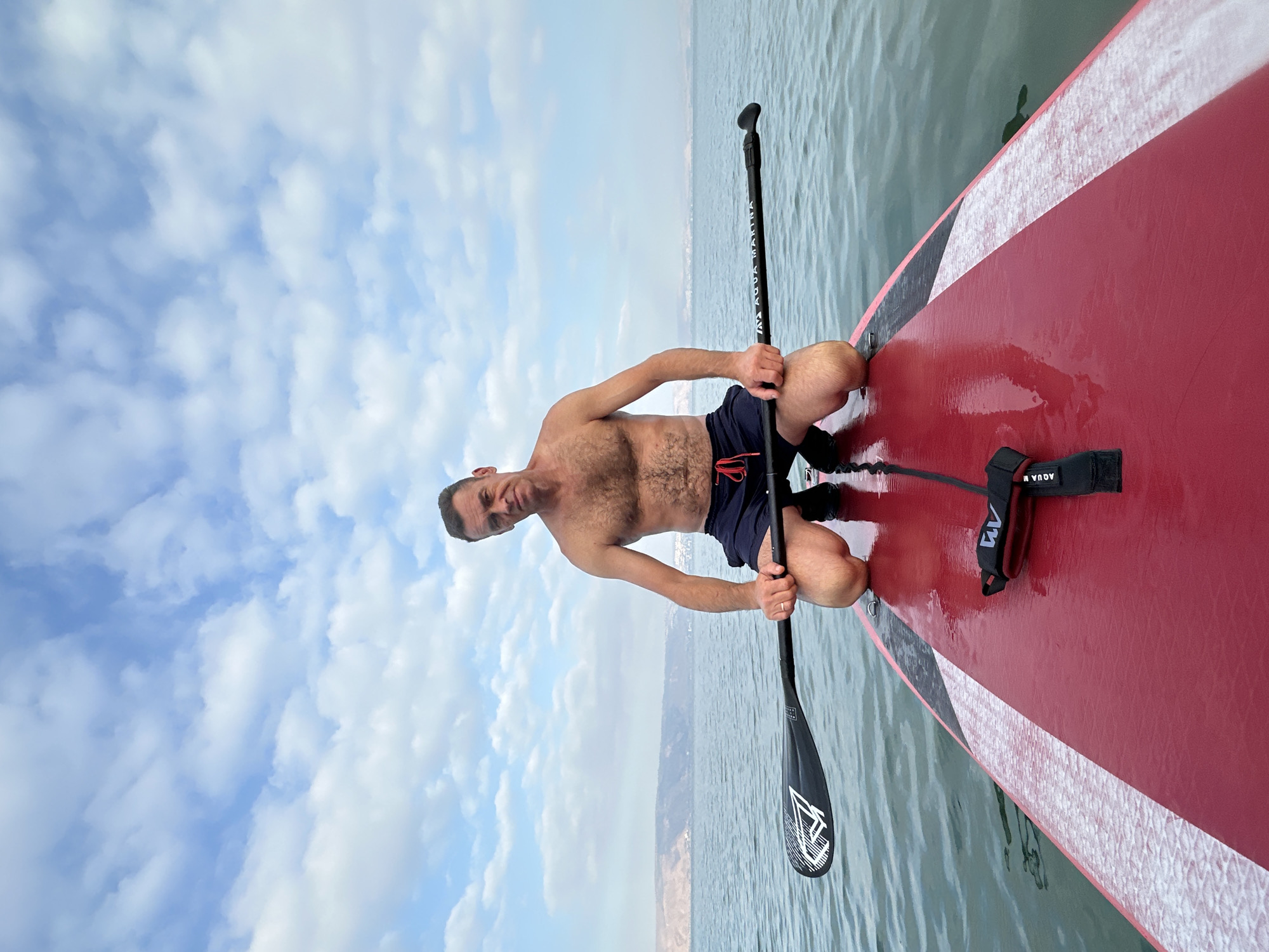 A shirtless man with short brown hair and a muscular build kneels on a red paddleboard. He is wearing dark blue swim trunks with a red drawstring. His hands rest on a black paddle that is positioned horizontally across the paddleboard. The man is looking directly at the camera with a neutral expression. The paddleboard is on calm, blue water. In the background, a hazy shoreline with hills and some buildings is visible under a cloudy sky with scattered white clouds. The scene appears to be set in the Sea of Galilee, located in the Emek HaYarden Regional Council, Israel.