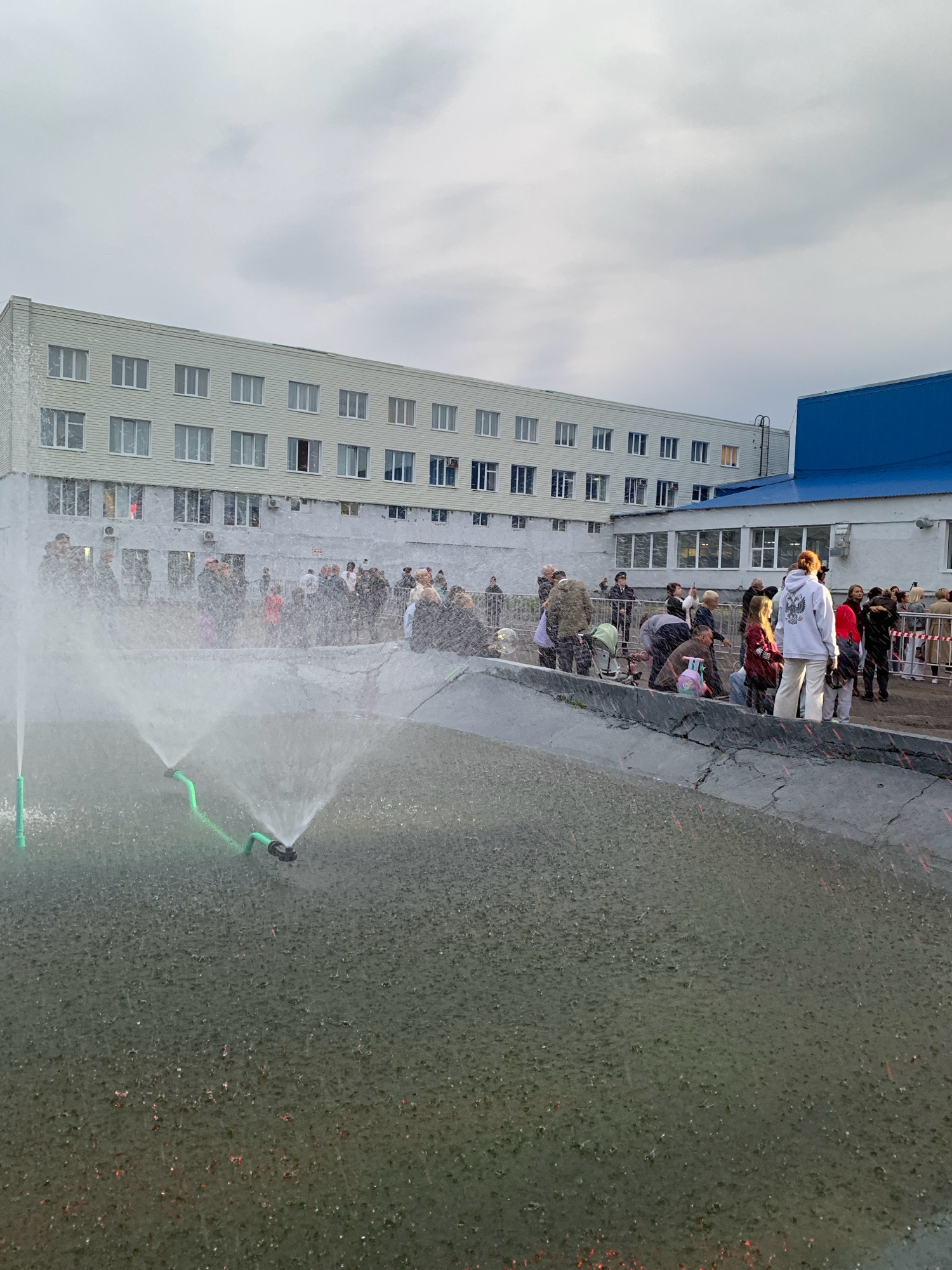 The image shows a group of people gathered outdoors in front of a light-colored, multi-story building, likely an administrative or educational facility. In the foreground, a water feature with two green sprinklers sprays water upwards, creating a mist. Some people are watching the water, while others appear to be interacting with each other. The building has numerous windows and appears to be part of a larger complex, as a blue-roofed structure is visible to the right. The sky is overcast, suggesting a cloudy day. The scene takes place in the city of Prokopyevsk, Russia.
