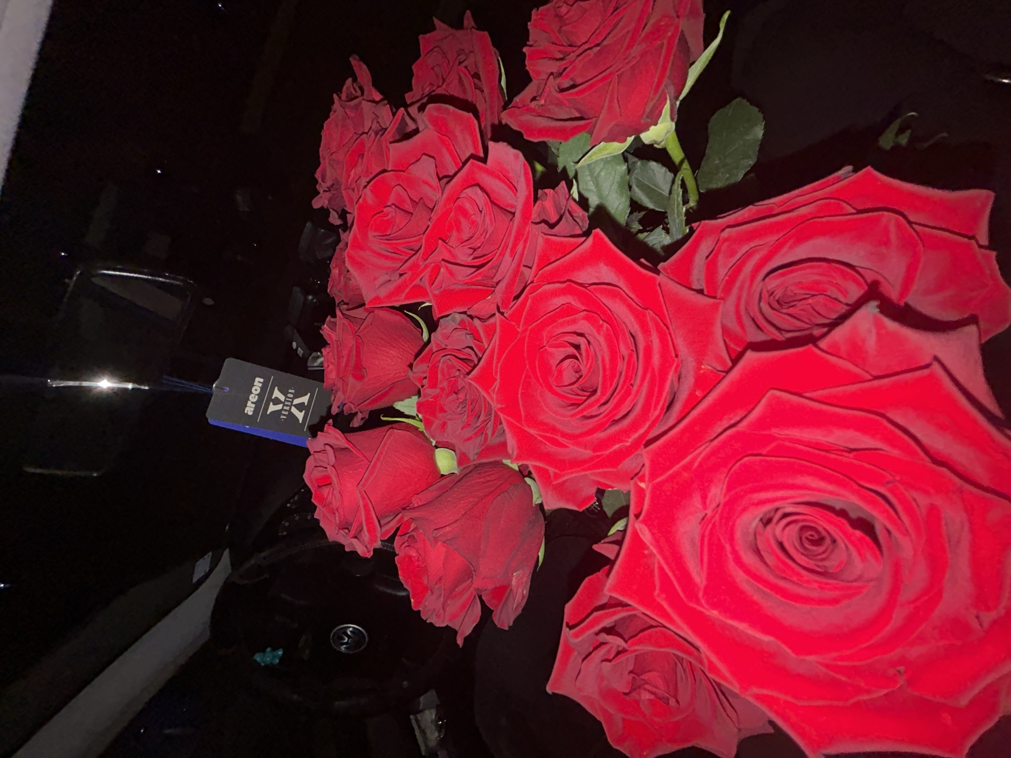 A close-up shot shows a bouquet of deep red roses resting on what appears to be the dashboard of a car. An air freshener with "areon" and "X-VERSION" printed on it hangs near the roses. The car interior is mostly black. The location is not identifiable as Zlatograd, Bulgaria from the content of this image.