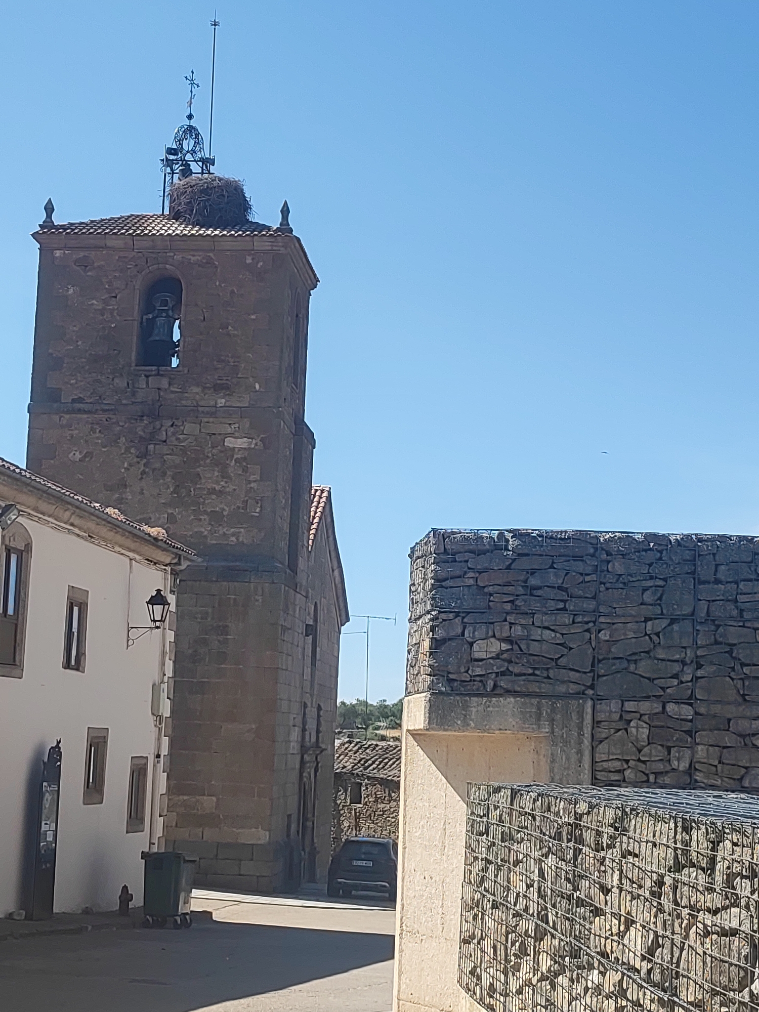 The image depicts a sunlit scene in the town of Sobradillo, Spain, focusing on a historic stone church tower. Atop the tower, a large stork's nest is prominently visible, with a weather vane and cross above it. A bell hangs within an arched opening of the tower. To the left, a white building with visible windows and a traditional street lamp stands adjacent to the church. In the foreground, a gabion wall, constructed with wire cages filled with stones, lines the street. A dark-colored car is parked on the street, and a green trash bin is situated near the white building. The sky is clear and blue. The architecture suggests a European setting, consistent with the identified location in Spain.