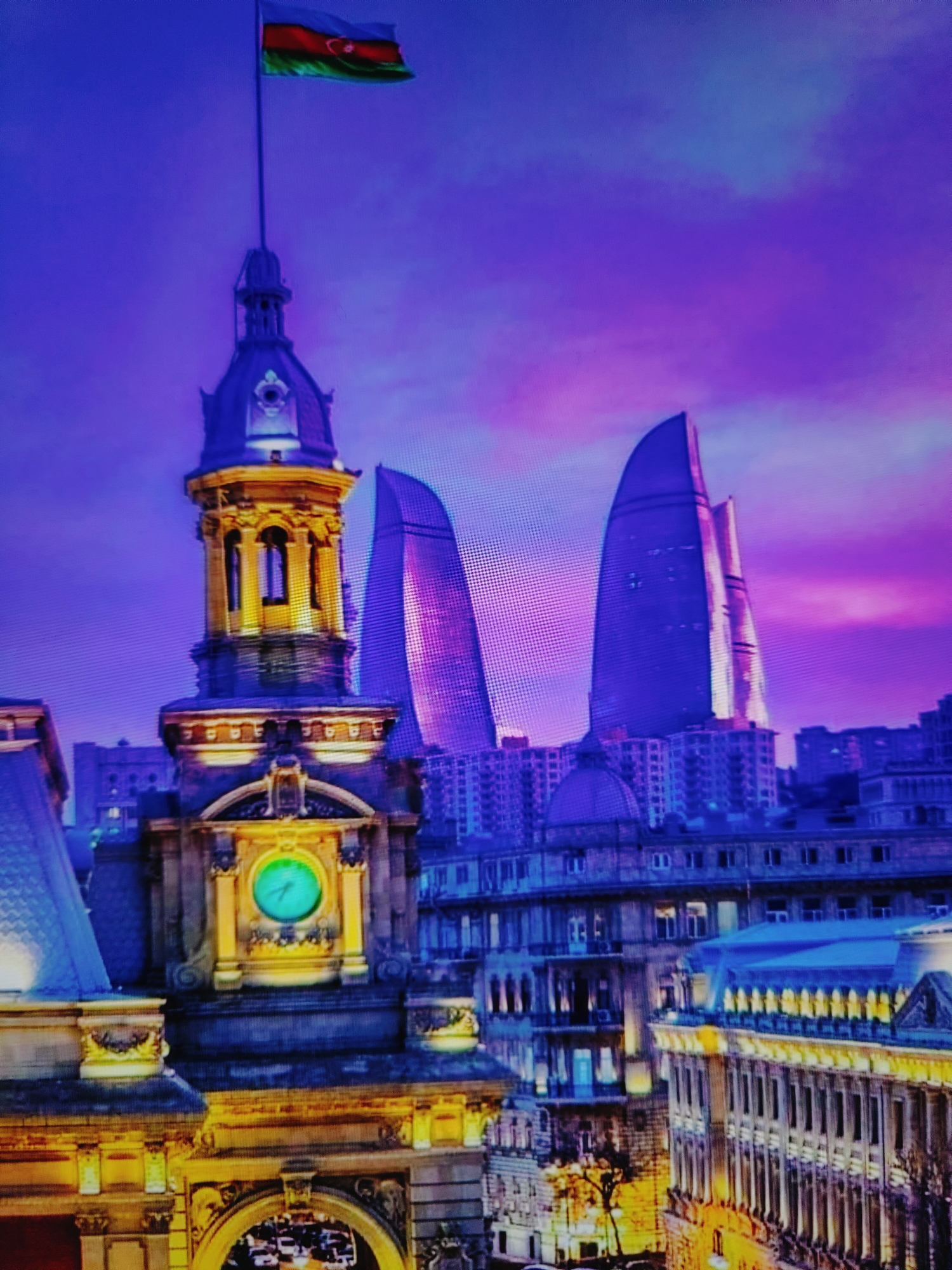 The image shows a view of Baku, Azerbaijan, at dusk or dawn. In the foreground, a historic building with a prominent clock tower is illuminated. The clock tower is adorned with golden architectural details and has a green clock face. Behind the historic building, two modern flame-shaped skyscrapers, part of the Flame Towers complex, dominate the skyline. An Azerbaijani flag flies from a flagpole atop the clock tower. The sky is a vibrant mix of purple and blue, indicating twilight. The surrounding cityscape consists of other buildings, some of which are also illuminated.