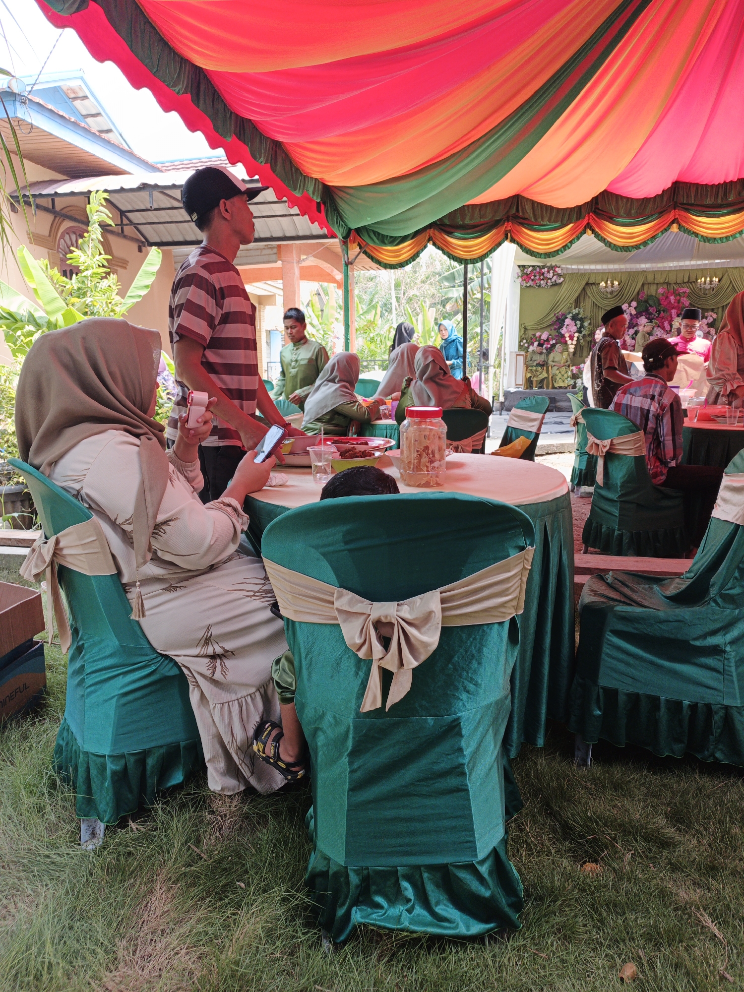In Pematang Duku, Indonesia, an outdoor gathering is taking place under a brightly colored tent with red, orange, and green fabric. Several round tables are set up, draped with green tablecloths and adorned with beige sashes tied in bows around the chairs. In the foreground, a woman wearing a light-colored headscarf and dress is seated, holding a smartphone and a small white camera in her hands. She is looking at her phone, which is resting on the table. To her right, another chair is occupied by a person whose lower legs and feet are visible, wearing sandals. In the background, more people are present, some standing and some seated at tables. One man in a striped shirt and a baseball cap stands near the foreground table, looking away from the camera. Other individuals are seen in the middle ground and further back, some wearing traditional clothing. The ground is covered in green grass. The overall scene suggests a celebratory or communal event.