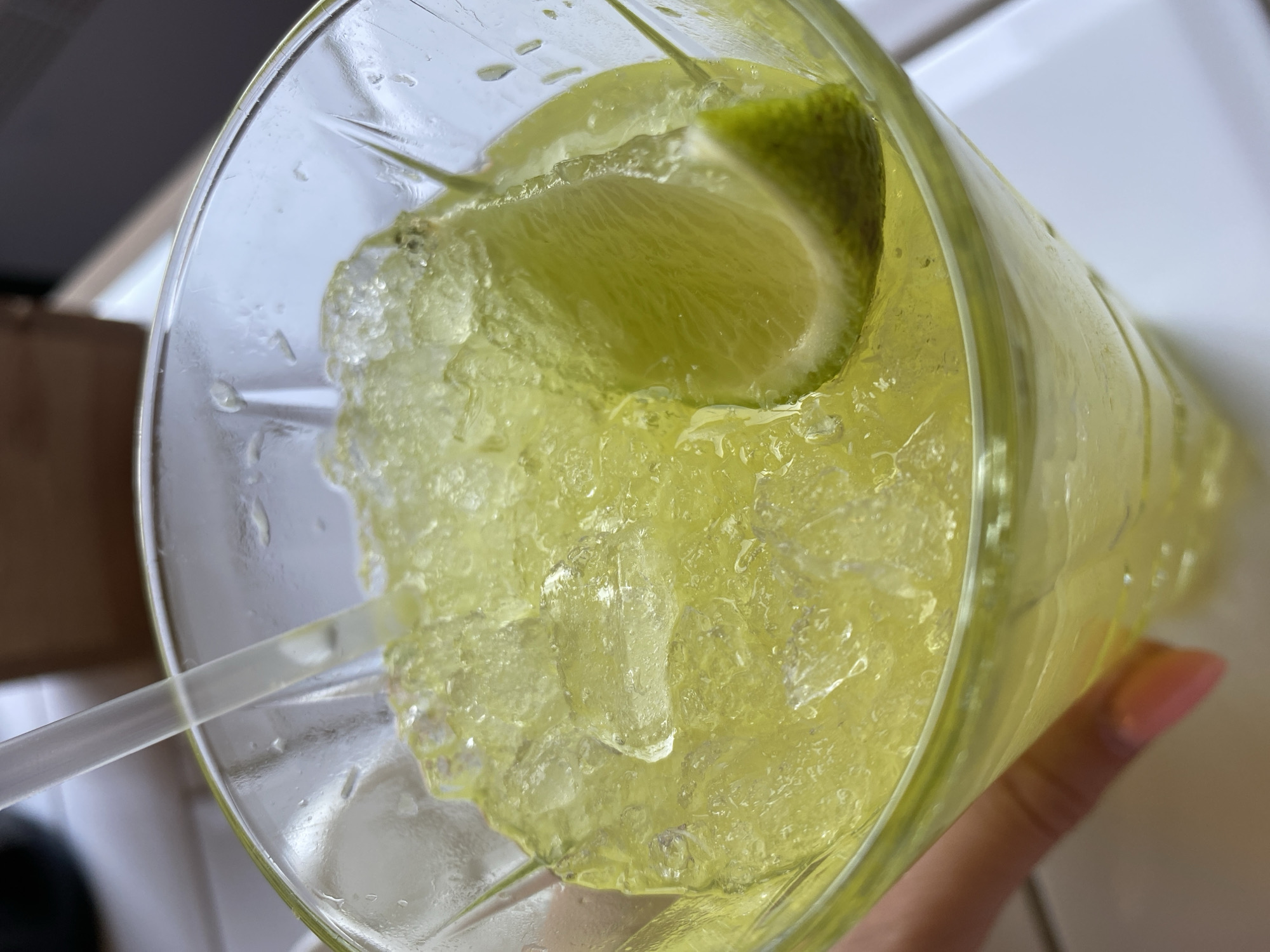 A close-up, high-angle shot shows a glass filled with crushed ice, a yellow-green liquid, and a lime wedge. A transparent straw is visible within the glass. The glass is held by a hand with a pink nail. The background includes a white countertop and a blurry dark gray object. There is no information indicating this image was taken in the city of Izhevsk, Russia.