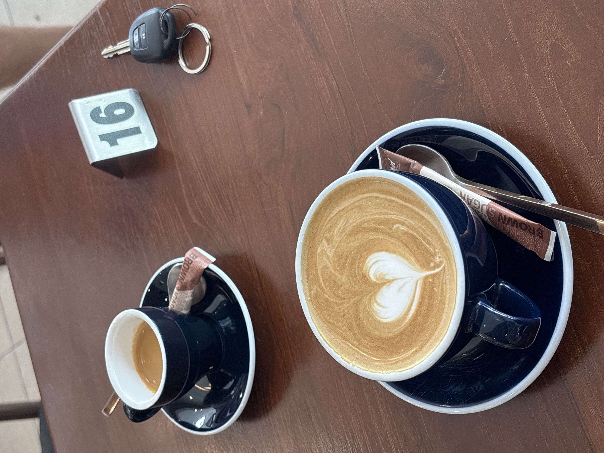 The image depicts a wooden table in what appears to be a cafe setting. Located on the table is a car key, a table number "16" sign, a small cup of espresso, and a larger cup of latte with a heart-shaped latte art. A brown sugar packet and a spoon are placed on the saucer of the latte. Given the mention of Choeng Thale, Thailand, it is inferred the image was taken in a cafe in that location. There are no people fully visible in the image, only a small part of an arm.