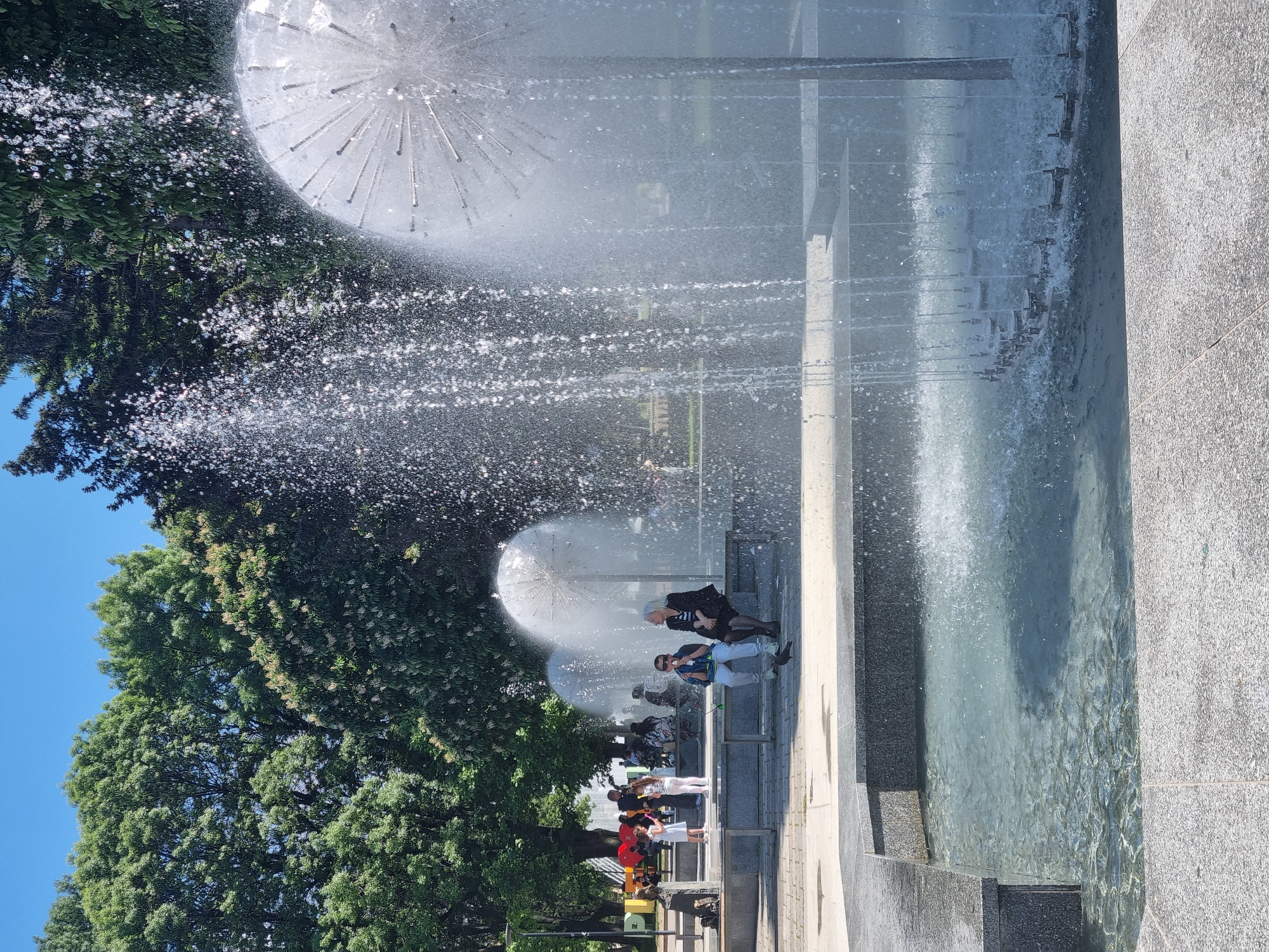 The image shows the Pleven Water Cascade, a fountain complex in Pleven, Bulgaria. The foreground features a water basin with jets spraying upwards. Several people are walking and sitting around the fountain. The fountains are spherical in shape and spray water in all directions. Trees are visible alongside the fountain. The sky is blue.
