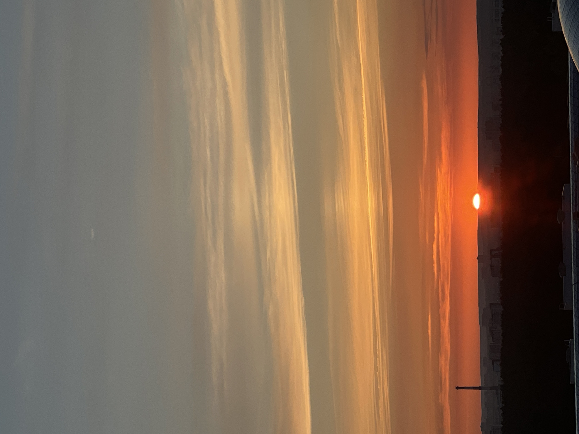 The image depicts a vibrant sunset over the city of Perm, Russia. The sky is a canvas of fiery oranges and soft yellows, transitioning to a muted gray at the top, with delicate wispy clouds stretching horizontally. In the foreground, a silhouetted cityscape lies beneath the dramatic sky, with a prominent industrial chimney standing tall on the left. The sun, a bright orange orb, is partially visible on the horizon, casting a warm glow. The foreground also shows the dark silhouette of trees and buildings, with hints of the urban landscape stretching out towards the sunset. The overall scene captures the serene beauty of a sunset in an industrial city.