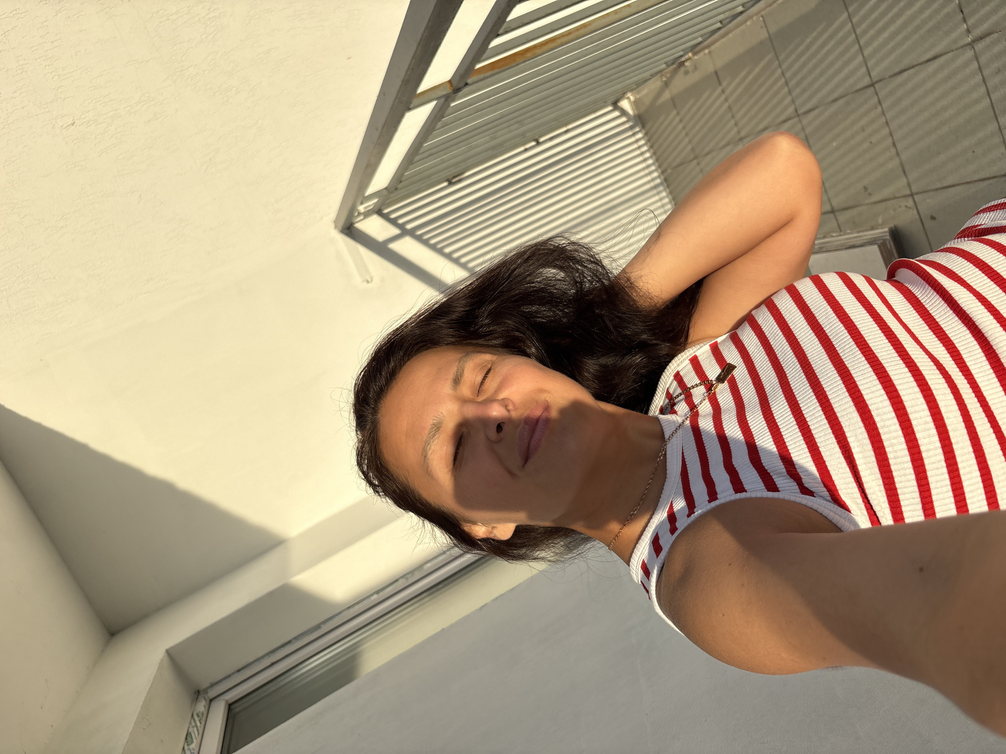 A woman with dark hair is lying down, with her eyes closed and her right arm behind her head. She is wearing a red and white striped top and a gold necklace. She appears relaxed and content, with sunlight illuminating her face. The background consists of a textured white wall and a railing with horizontal slats, casting striped shadows on the wall. The scene is outdoors, possibly on a balcony or patio, given the railing and outdoor lighting. The strong sunlight suggests it is daytime, likely morning or late afternoon due to the angle of the shadows. The overall mood is peaceful and serene.